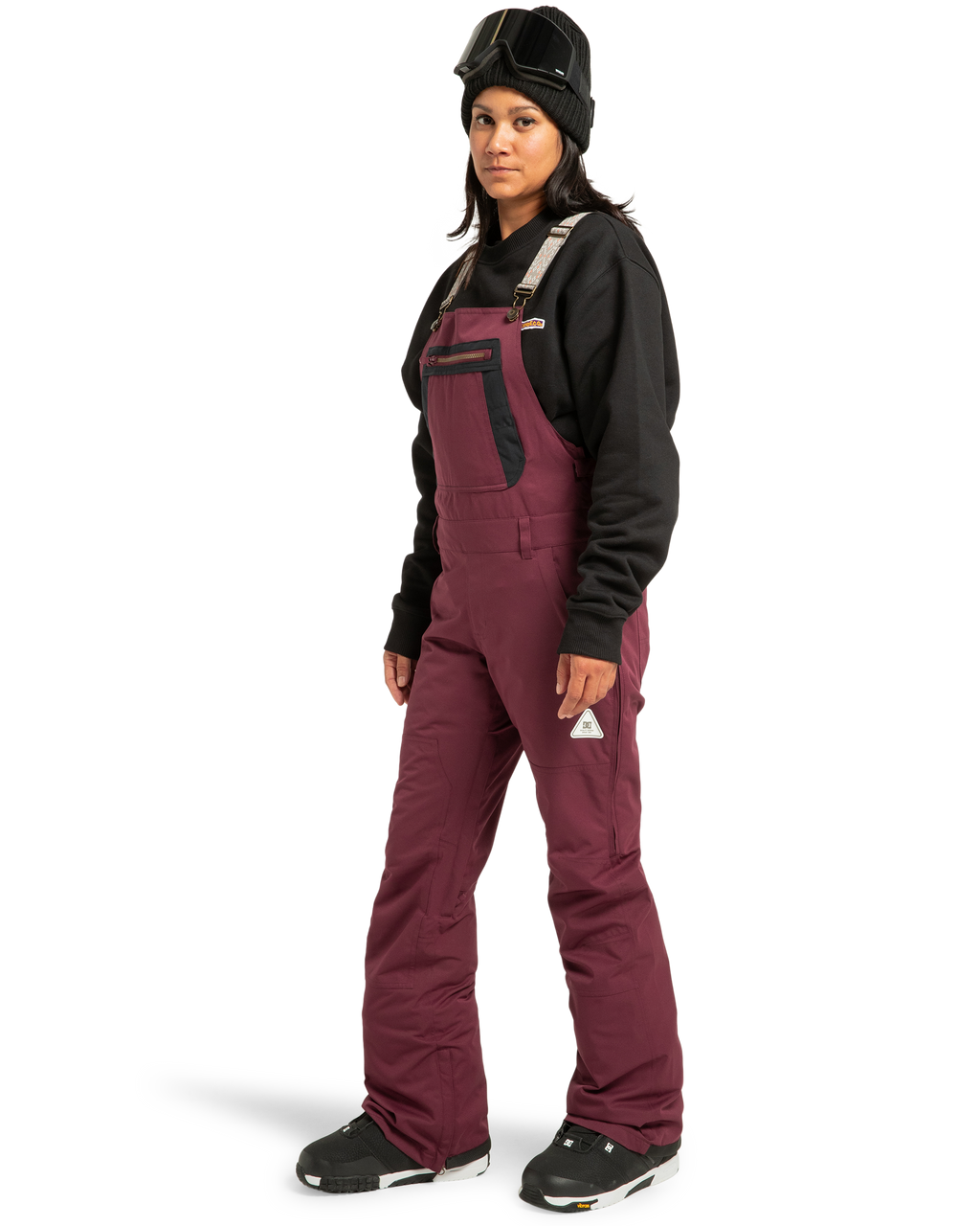 A person wearing a black beanie, ski goggles on their head, a black sweatshirt, and DC Shoes Women’s Crusade Shell Bib overalls in maroon with black boots, standing against a plain white background.