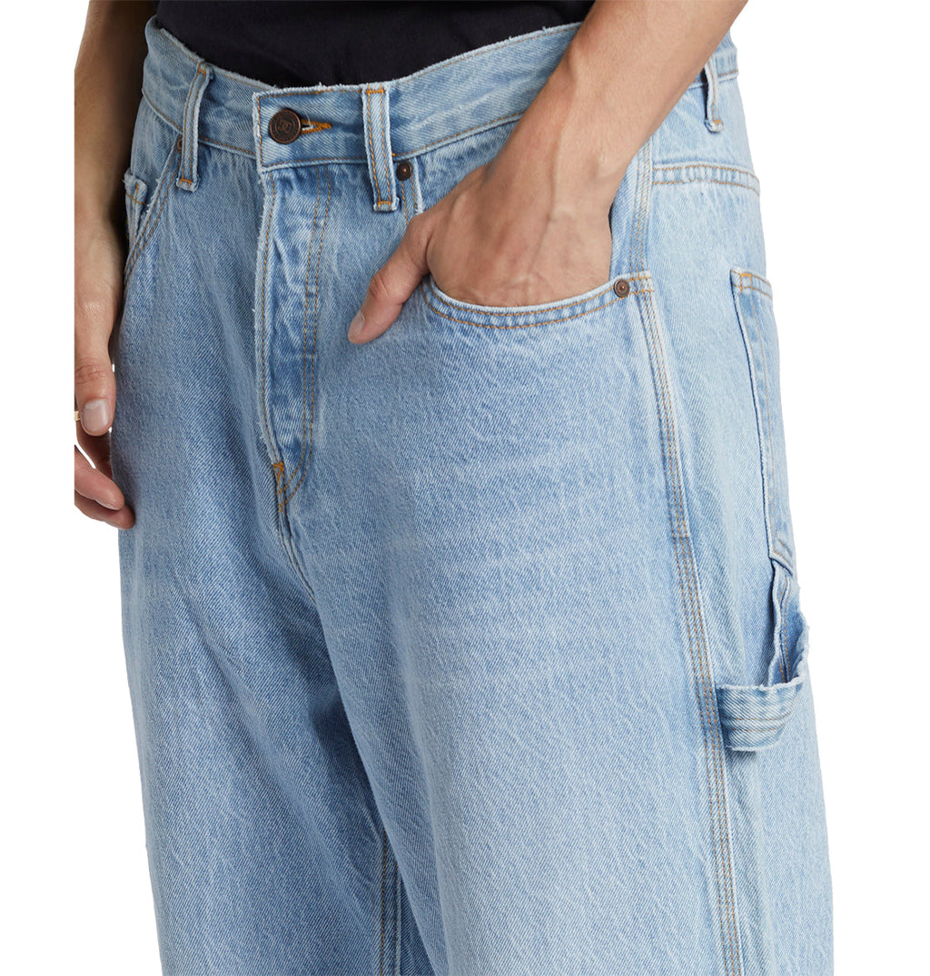A person wearing DC Shoes Men’s Worker Baggy Carpenter Ril Jeans in light blue denim with one hand tucked into the front pocket. These jeans feature a relaxed, baggy fit, visible stitching, and embody a laid-back vibe inspired by classic workwear style.