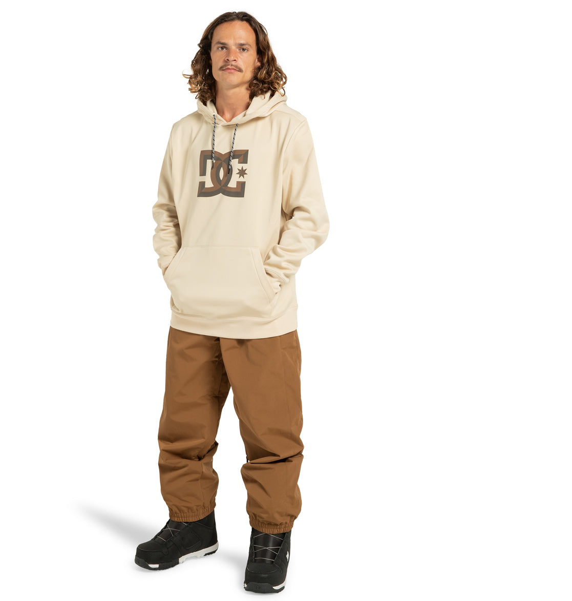 A man with wavy brown hair wears a beige DC Shoes Unisex Snowstar Hoodie, crafted from polyester bonded fleece, teamed with brown pants and black snow boots. Standing with his hands in the hoodie pocket against a plain white background, he showcases the weather defense style and comfort of the DC Shoes Snowstar Hoodie.