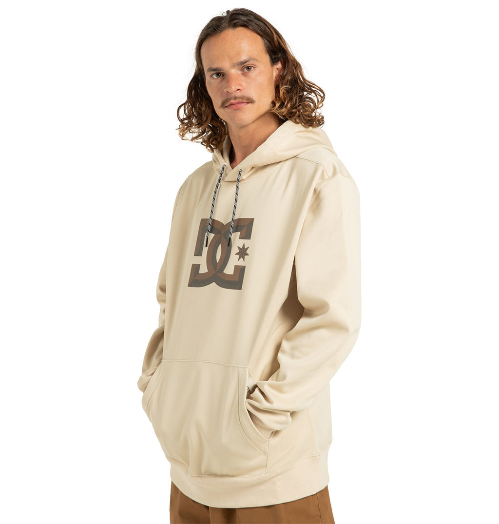 A man with long wavy hair wearing the DC Shoes Unisex Snowstar Hoodie—crafted from beige weather defense polyester bonded fleece and showcasing a large DC Shoes logo—paired with tan pants, stands with his hands in the hoodie pocket and looks at the camera against a white background.