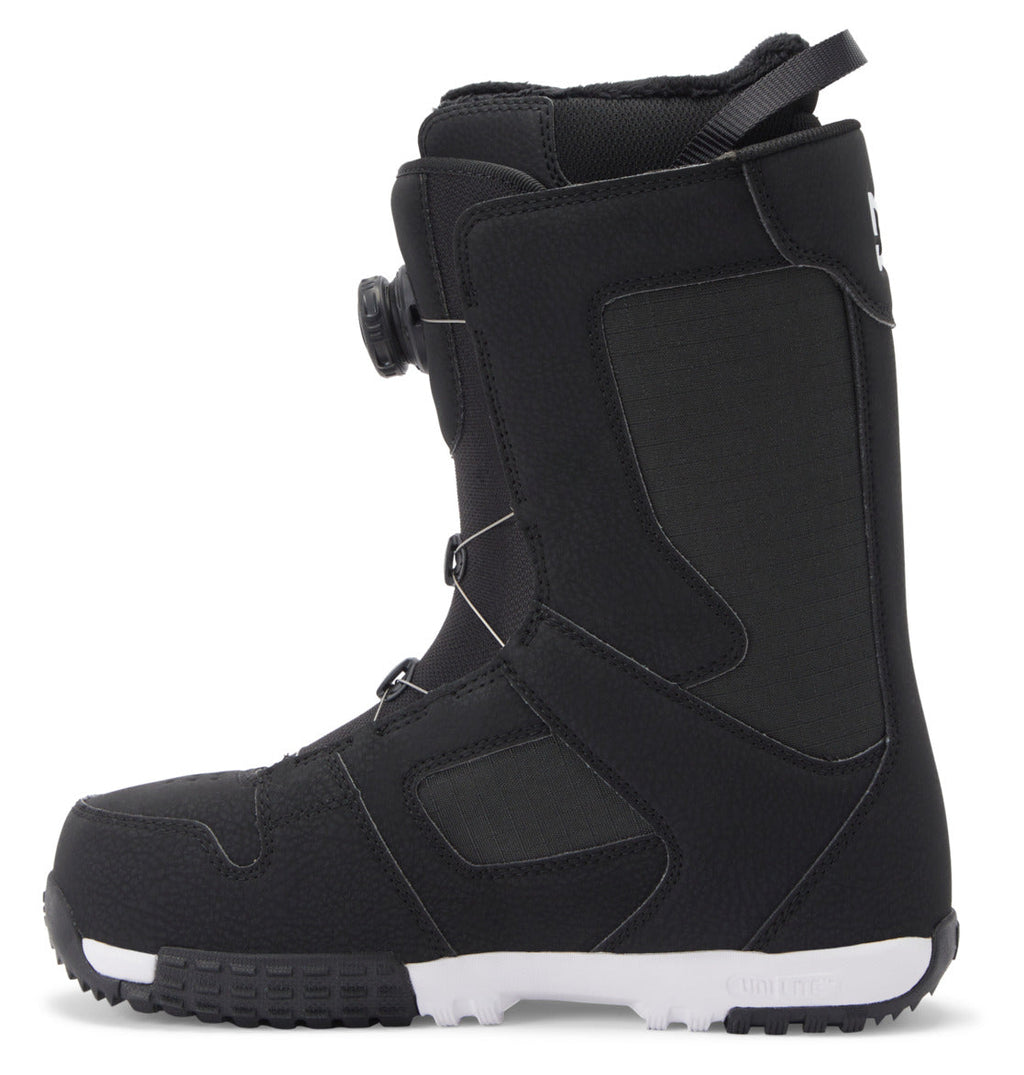 A black DC Shoes Men’s Phase Pro BOA® snowboard boot with textured material, a white sole, and a side dial lacing system. This boot features visible stitching, PrimaLoft insulation for added warmth, and a pull loop at the back.