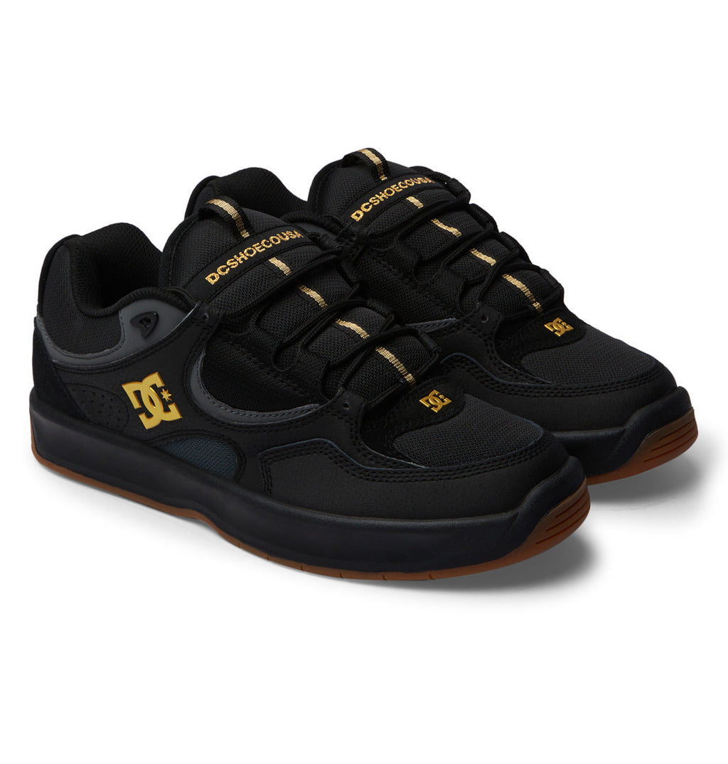 A pair of DC Shoes Men’s Kalynx Zero sneakers in black with gold accents, featuring black laces and brown rubber soles, displayed on a white background.