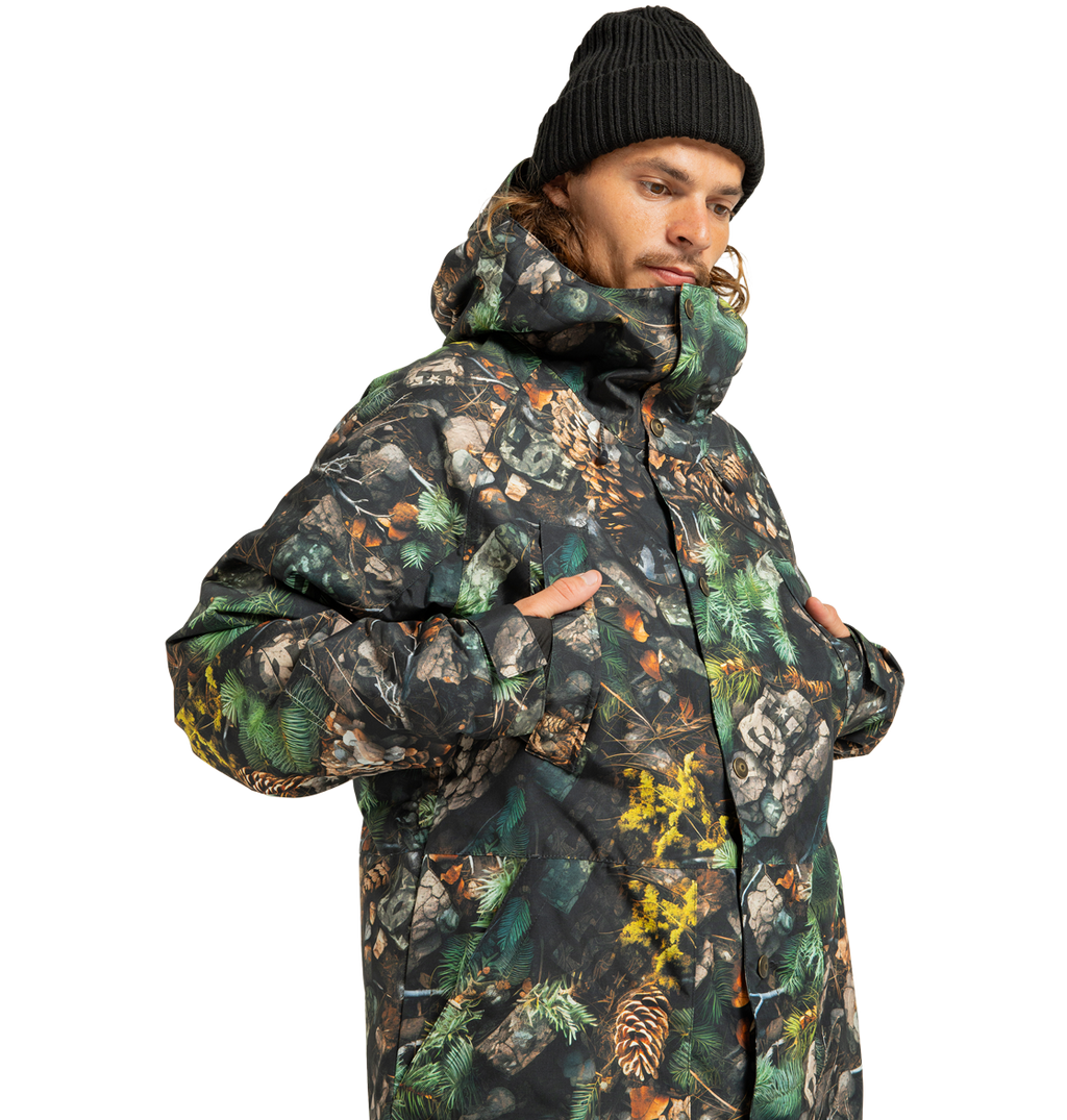 A person wearing a black beanie and the DC Shoes Men’s Tundra Insulated Jacket—an eco-friendly, camouflage-patterned men’s jacket with a hood, featuring tree branches and pine cones—stands with hands in pockets against a white background.