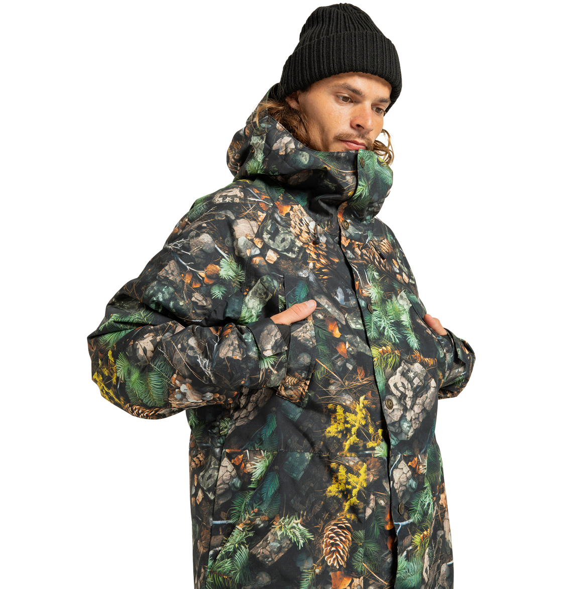 A person wearing a black beanie and the DC Shoes Men’s Tundra Insulated Jacket—an eco-friendly, camouflage-patterned men’s jacket with a hood, featuring tree branches and pine cones—stands with hands in pockets against a white background.