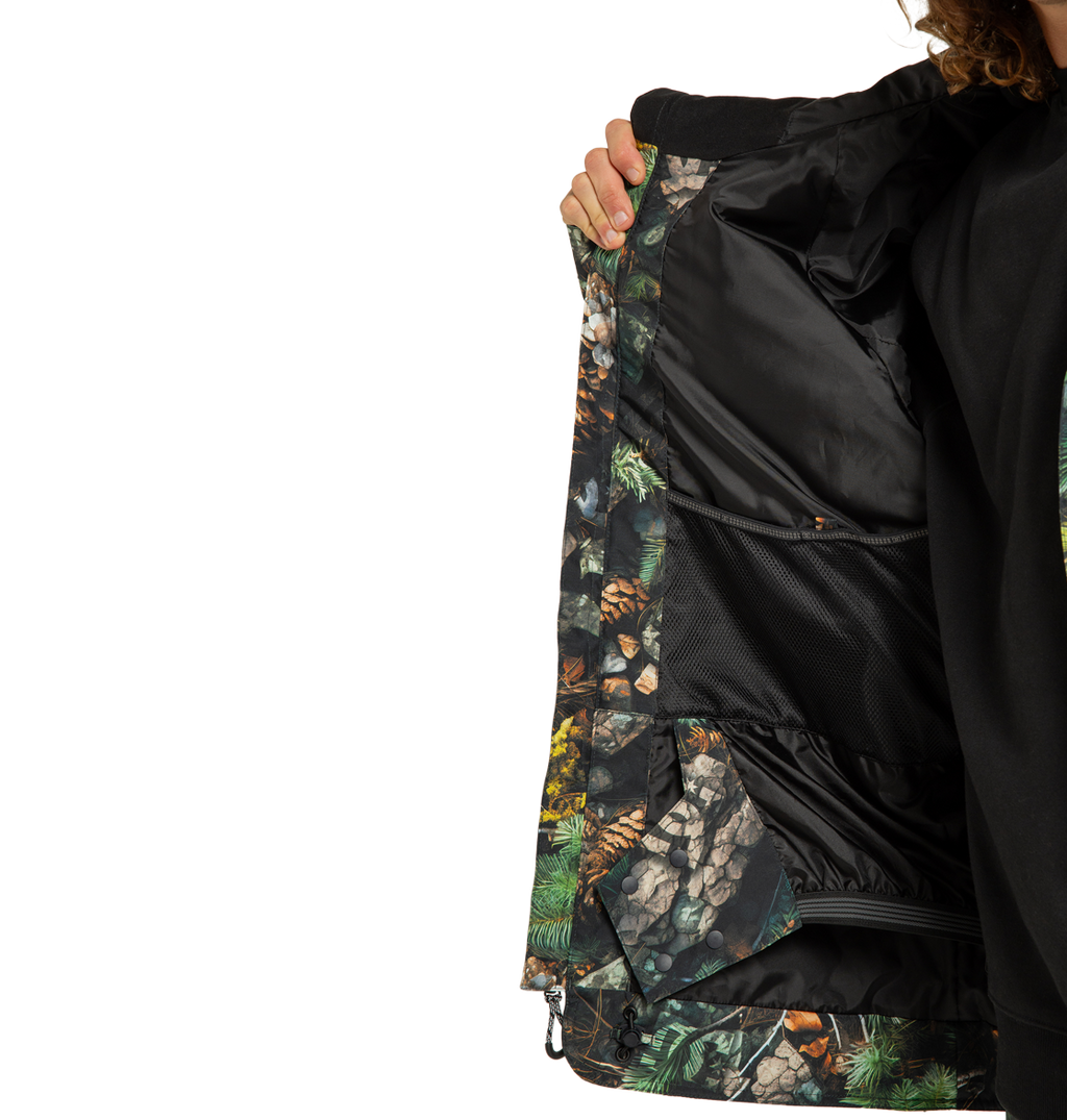 A person opens the DC Shoes Men’s Tundra Insulated Jacket to reveal its black mesh inner pocket and a lining adorned with a nature-themed print featuring pinecones, rocks, and greenery. Only part of the individual is visible.