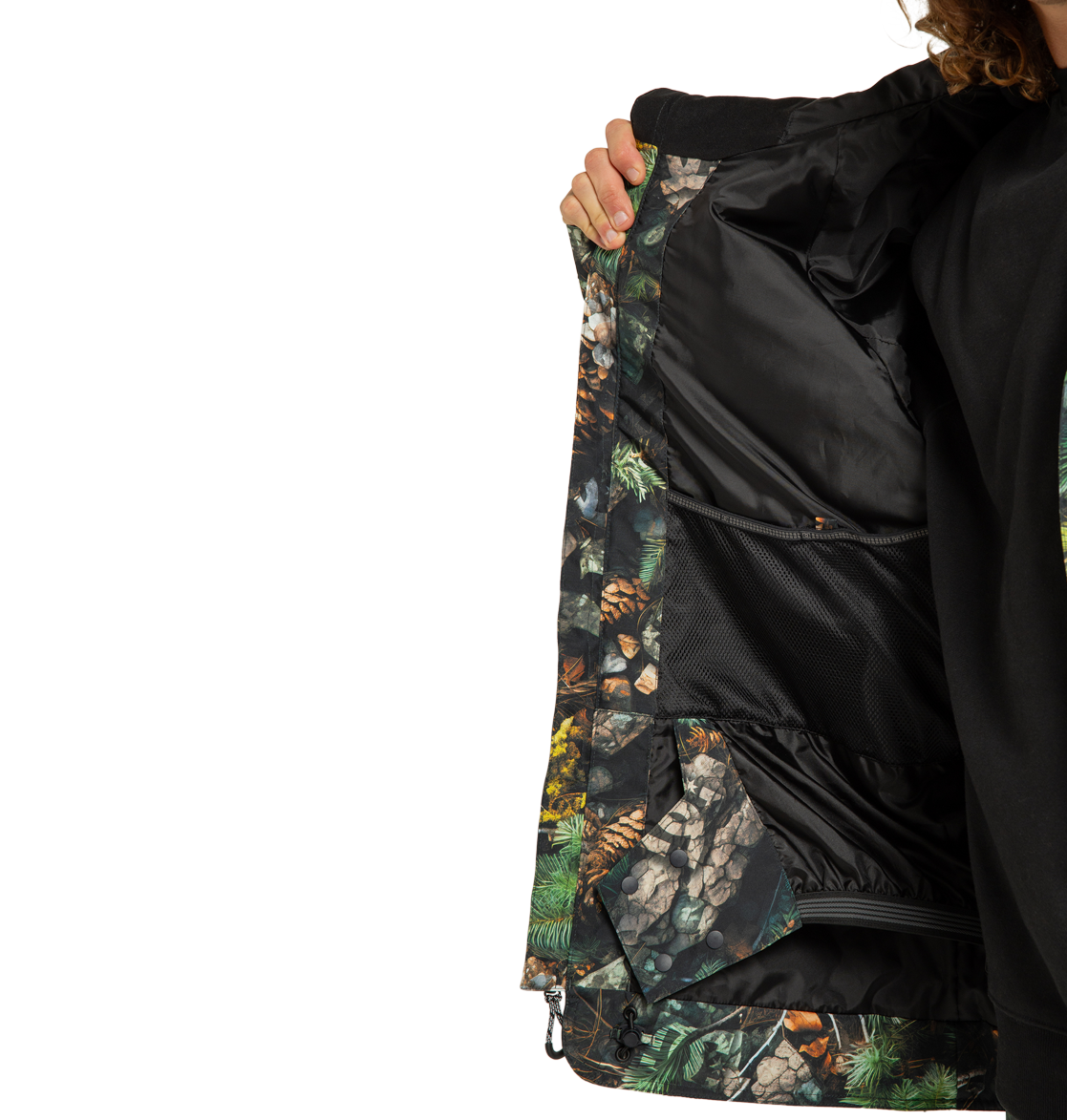A person opens the DC Shoes Men’s Tundra Insulated Jacket to reveal its black mesh inner pocket and a lining adorned with a nature-themed print featuring pinecones, rocks, and greenery. Only part of the individual is visible.