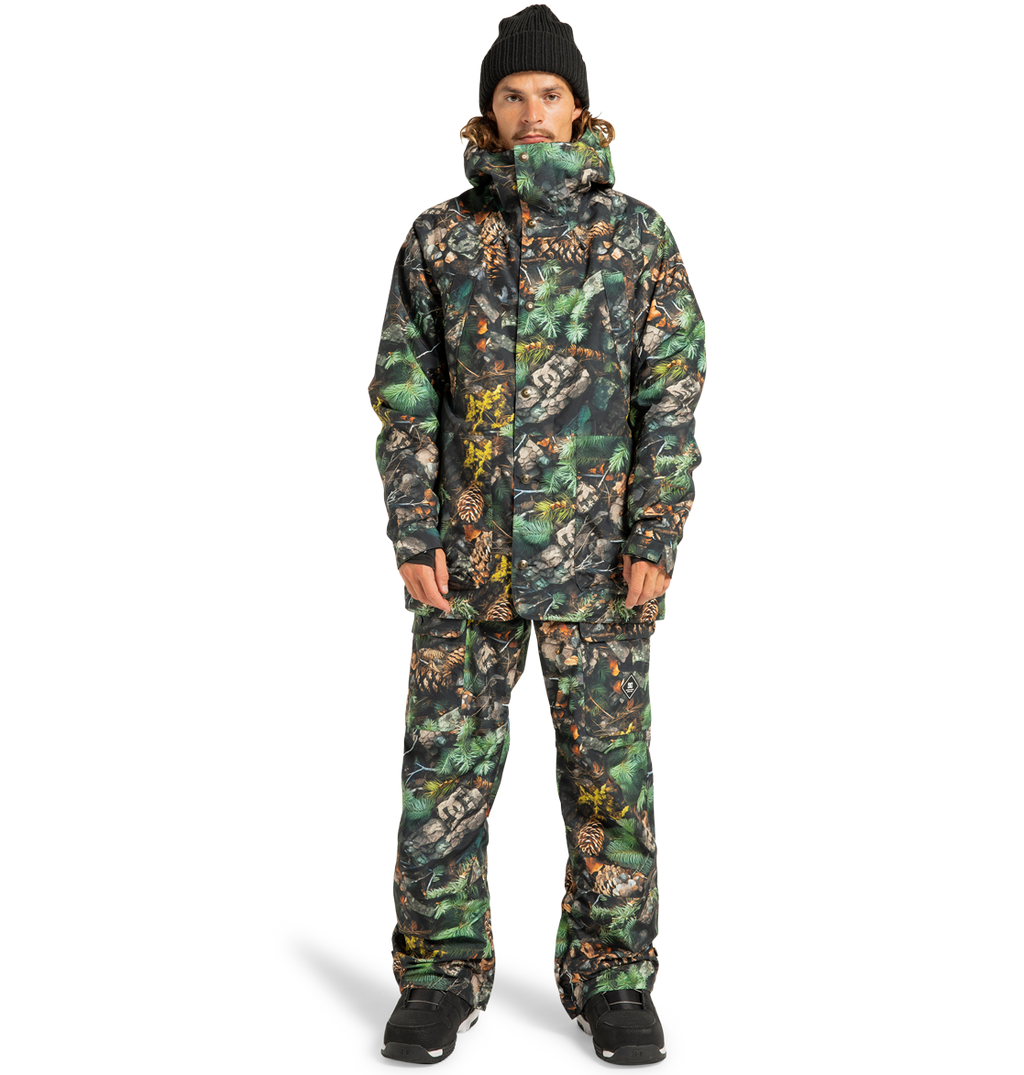 A person is standing facing forward against a plain white background, wearing a matching camouflage-patterned DC Shoes Men’s Tundra Insulated Jacket and pants, paired with a black beanie and black boots.