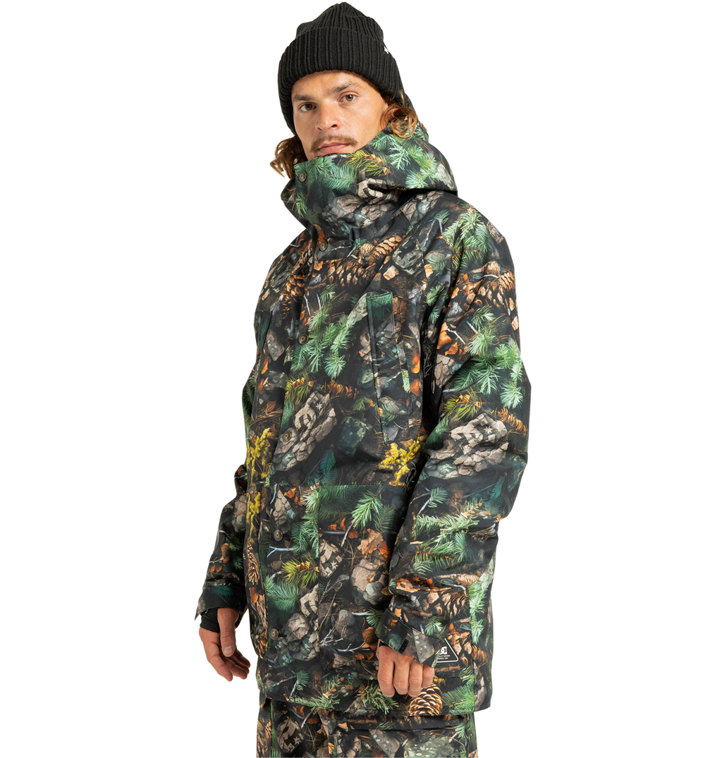 A person wearing a black beanie and the DC Shoes Men’s Tundra Insulated Jacket, featuring an eco-friendly green, brown, and black camouflage pattern, along with matching pants, stands and looks slightly to the side against a plain white background.
