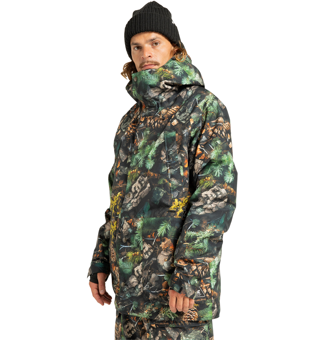 A person wearing a black beanie and the DC Shoes Men’s Tundra Insulated Jacket, featuring an eco-friendly green, brown, and black camouflage pattern, along with matching pants, stands and looks slightly to the side against a plain white background.