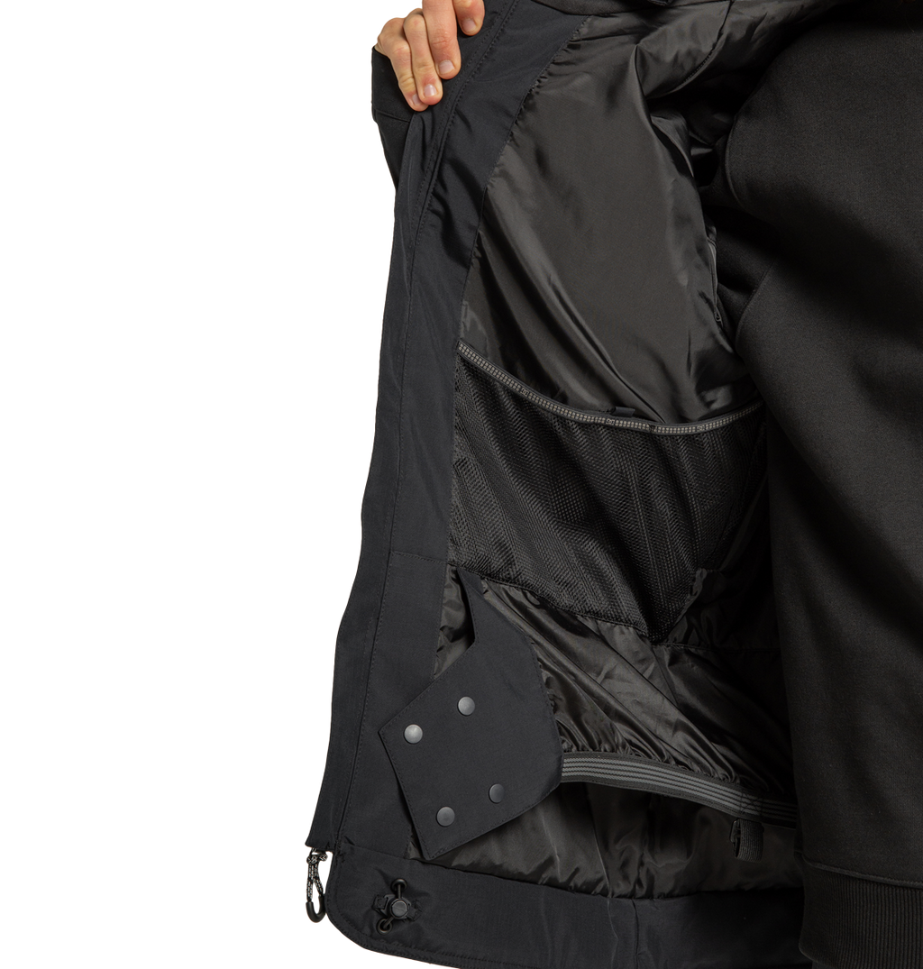 A person holds open a DC Shoes Men’s Tundra Insulated Jacket in black, revealing the inside lining, mesh pockets, and snap buttons. Only the person’s hand and part of their torso are visible.