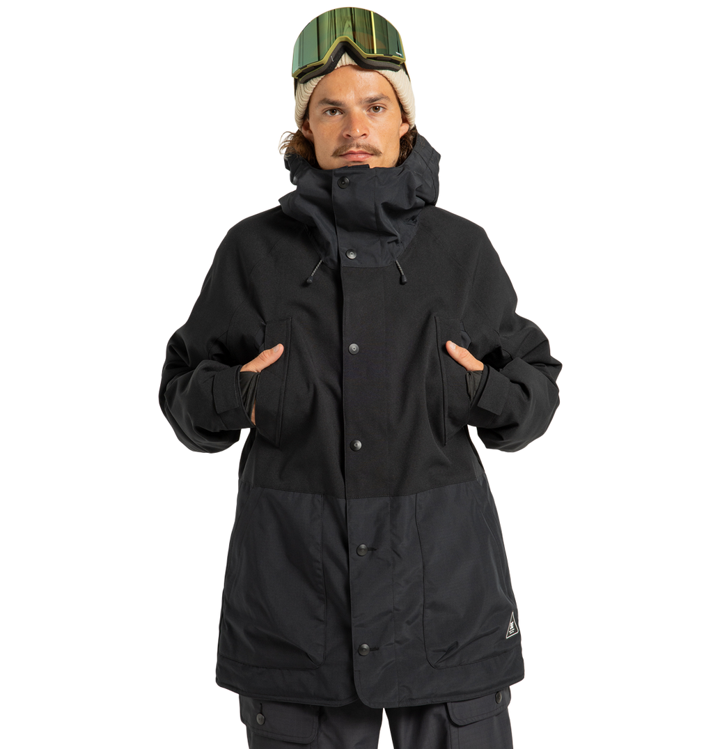 A person wearing the DC Shoes Men’s Tundra Insulated Jacket in black, paired with black pants and ski goggles, stands facing forward with hands in the jacket’s chest-high pockets against a plain white background.