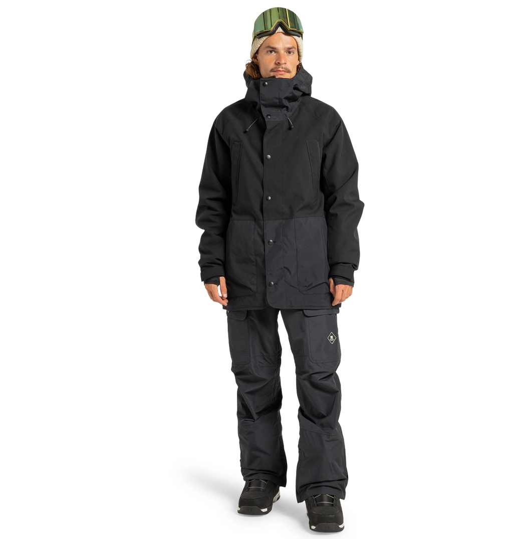 A person is wearing the DC Shoes Men’s Tundra Insulated Jacket and pants, black boots, and green ski goggles, standing against a plain white background. The insulated jacket offers exceptional warmth while adding a stylish edge to the overall look.