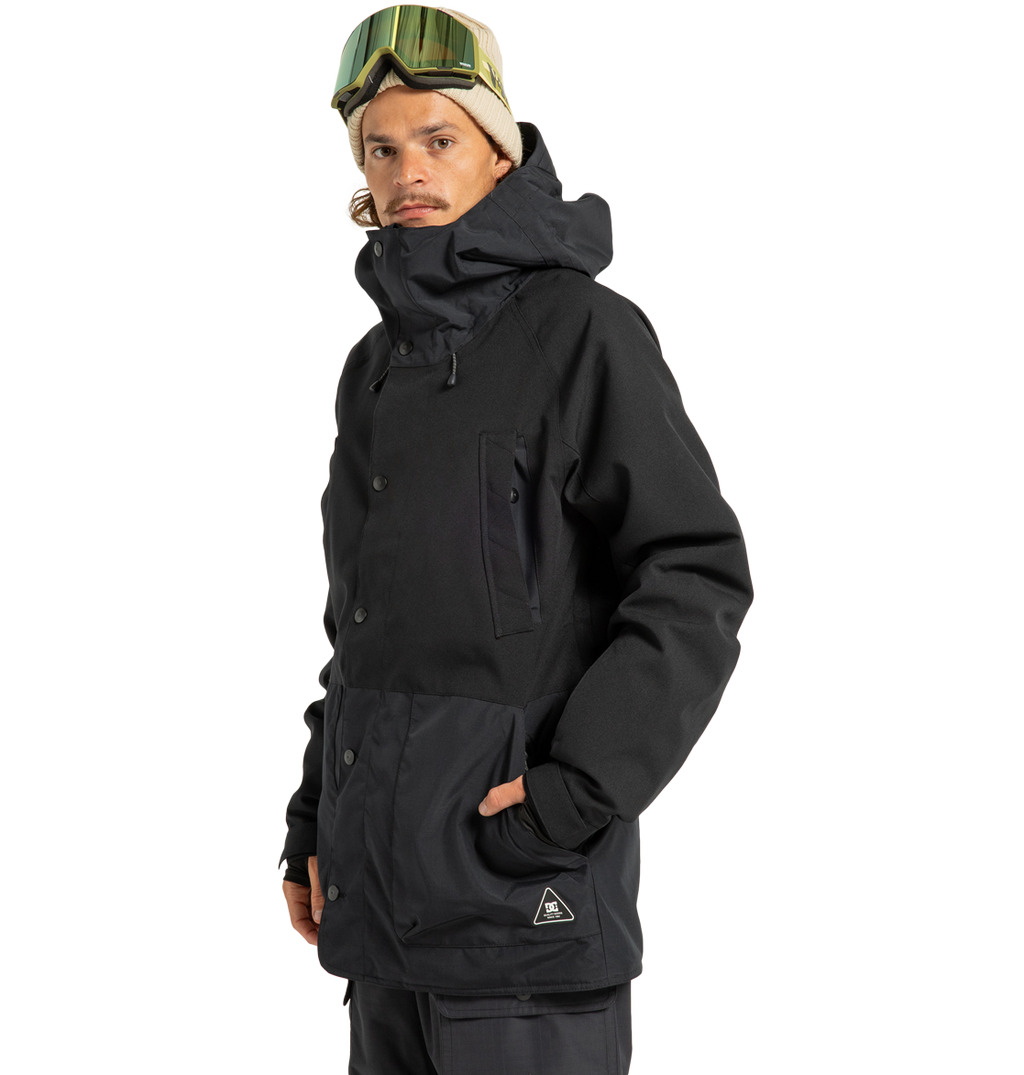 A person wearing the DC Shoes Men’s Tundra Insulated Jacket in black, paired with dark pants, a beige beanie, and ski goggles stands with one hand in their pocket against a plain white background.