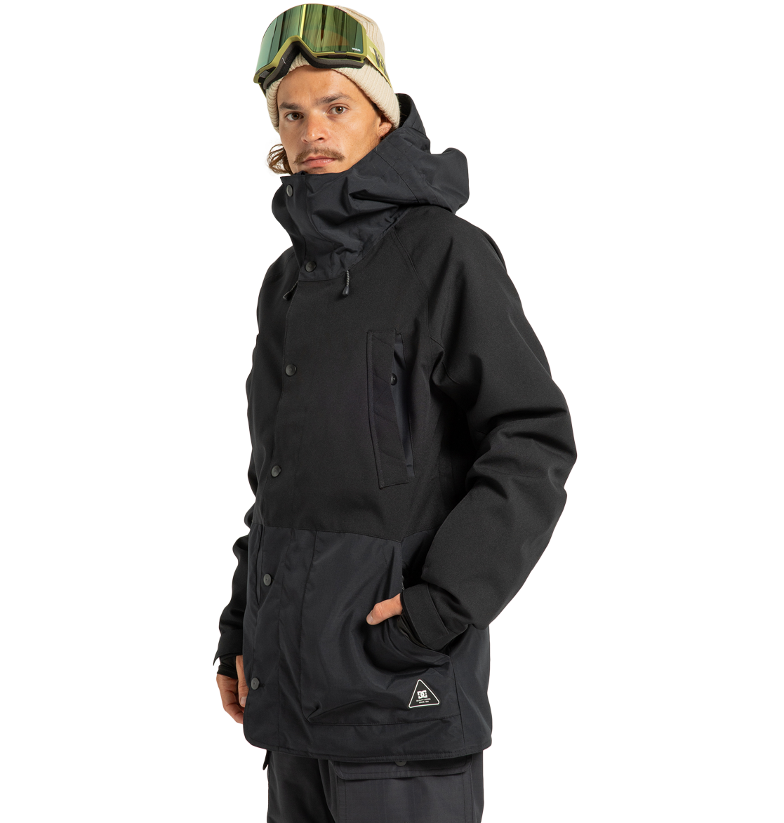 A person wearing the DC Shoes Men’s Tundra Insulated Jacket in black, paired with dark pants, a beige beanie, and ski goggles stands with one hand in their pocket against a plain white background.
