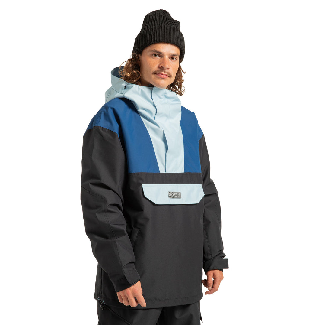 A person wearing a black beanie and the DC Shoes Men’s DC-43 Anorak Jacket in a color-blocked blue, light blue, and black design stands facing the camera with a neutral expression against a white background.