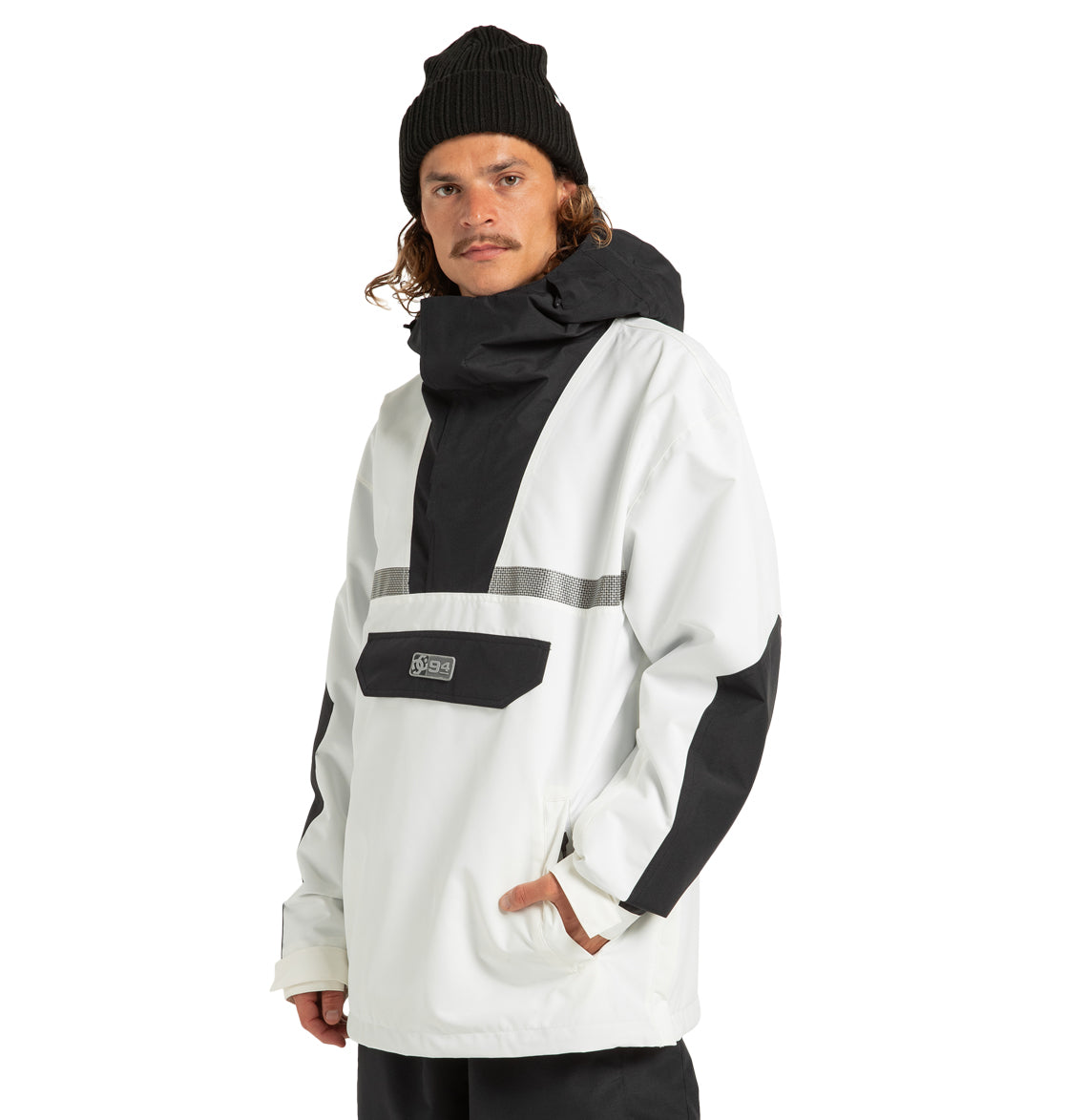 A man wearing a black beanie and the DC Shoes Mens’ DC-43 Anorak Jacket, designed in white and black with a front pocket, stands facing forward. This lightweight jacket features a high collar and a reflective stripe across the chest, combining both style and comfort.
