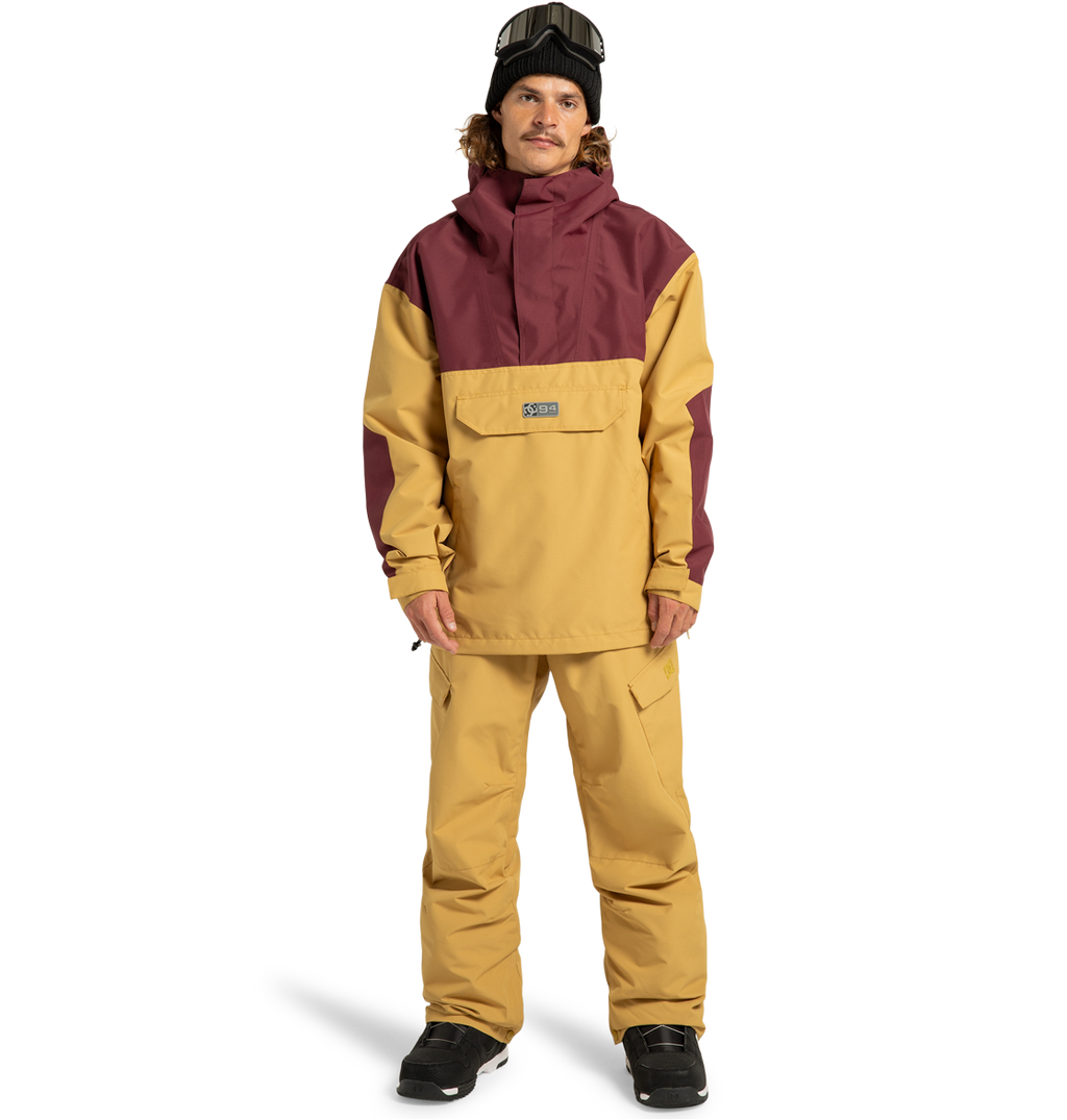 A person wearing the men’s DC-43 Anorak Shell Jacket by DC Shoes in yellow and maroon, paired with matching yellow snow pants, black gloves, black boots, a black beanie, and ski goggles stands against a plain white background.