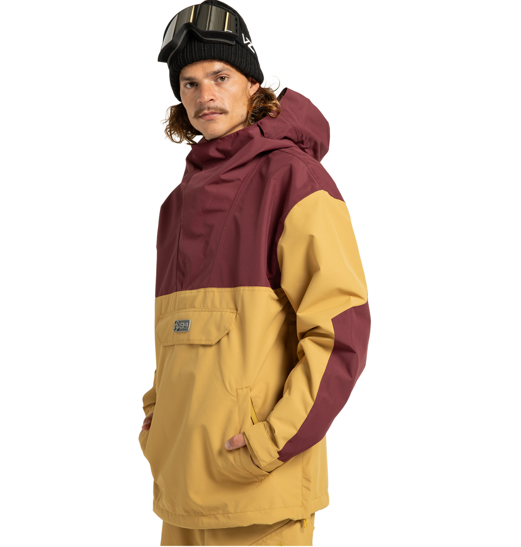 Wearing the Men’s DC-43 Anorak Shell Jacket by DC Shoes—a two-tone maroon and tan shell crafted from recycled polyester for enhanced sustainability—a person pairs it with tan pants, a black beanie, and ski goggles on their head. With hands in their pockets, they stand facing the camera.