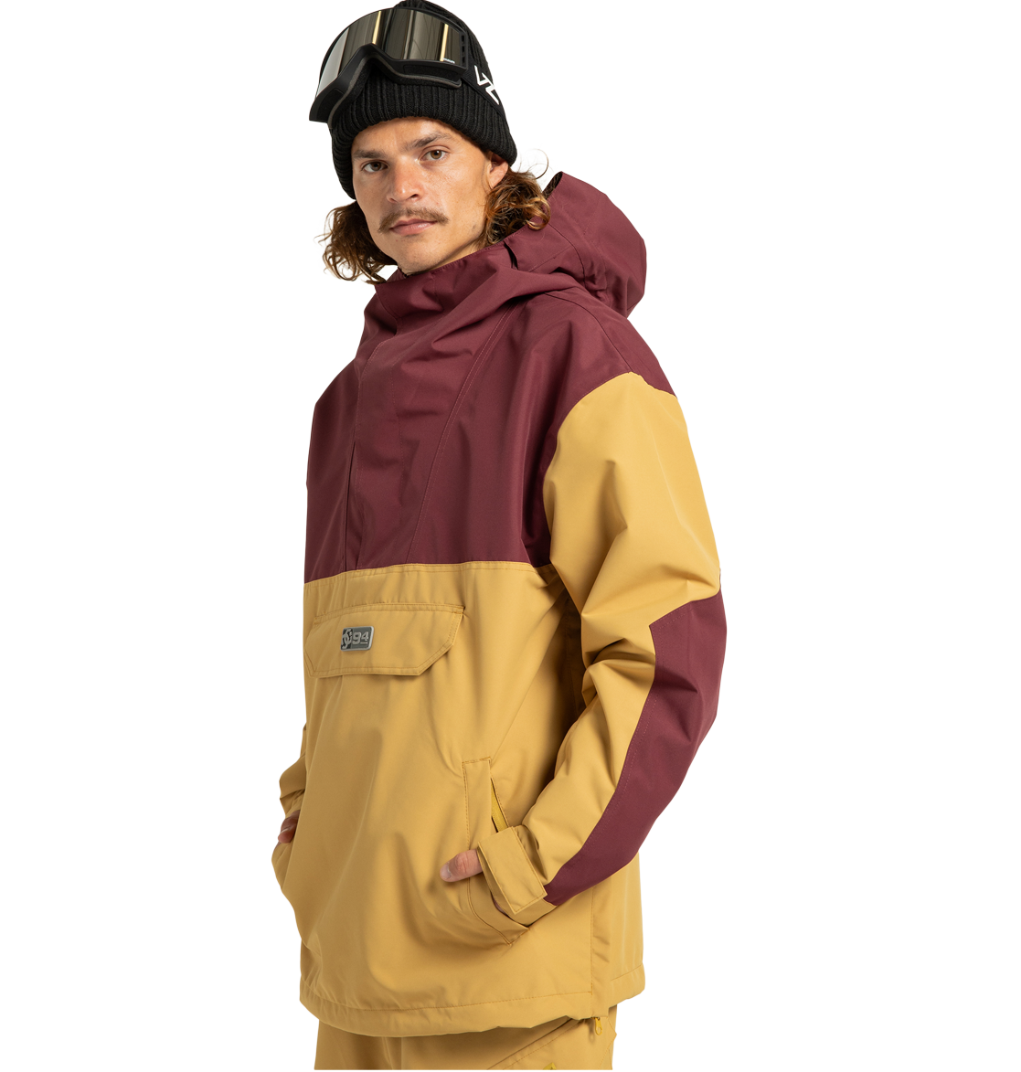 Wearing the Men’s DC-43 Anorak Shell Jacket by DC Shoes—a two-tone maroon and tan shell crafted from recycled polyester for enhanced sustainability—a person pairs it with tan pants, a black beanie, and ski goggles on their head. With hands in their pockets, they stand facing the camera.
