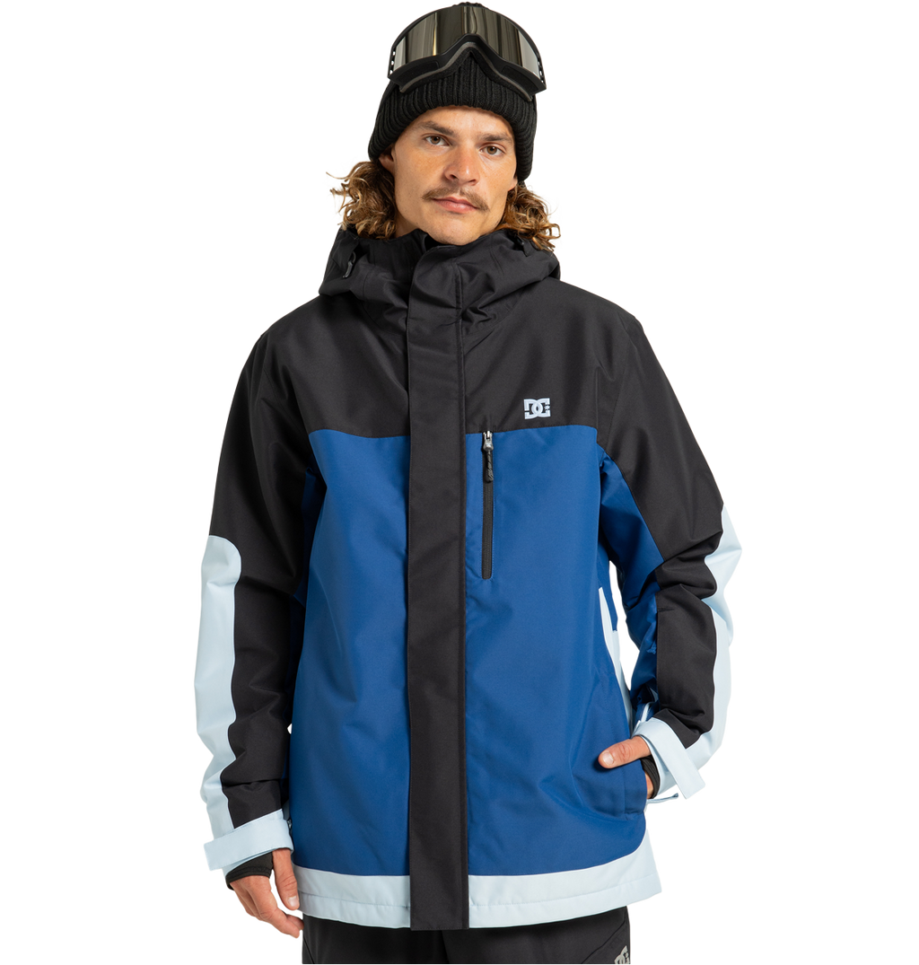 A man wearing the DC Shoes Men’s Defiant Insulated Jacket—crafted from recycled polyester in blue, black, and light blue with Weather Defense 10 technology—stands against a white background with one hand in his pocket, accessorized with a black beanie and ski goggles.