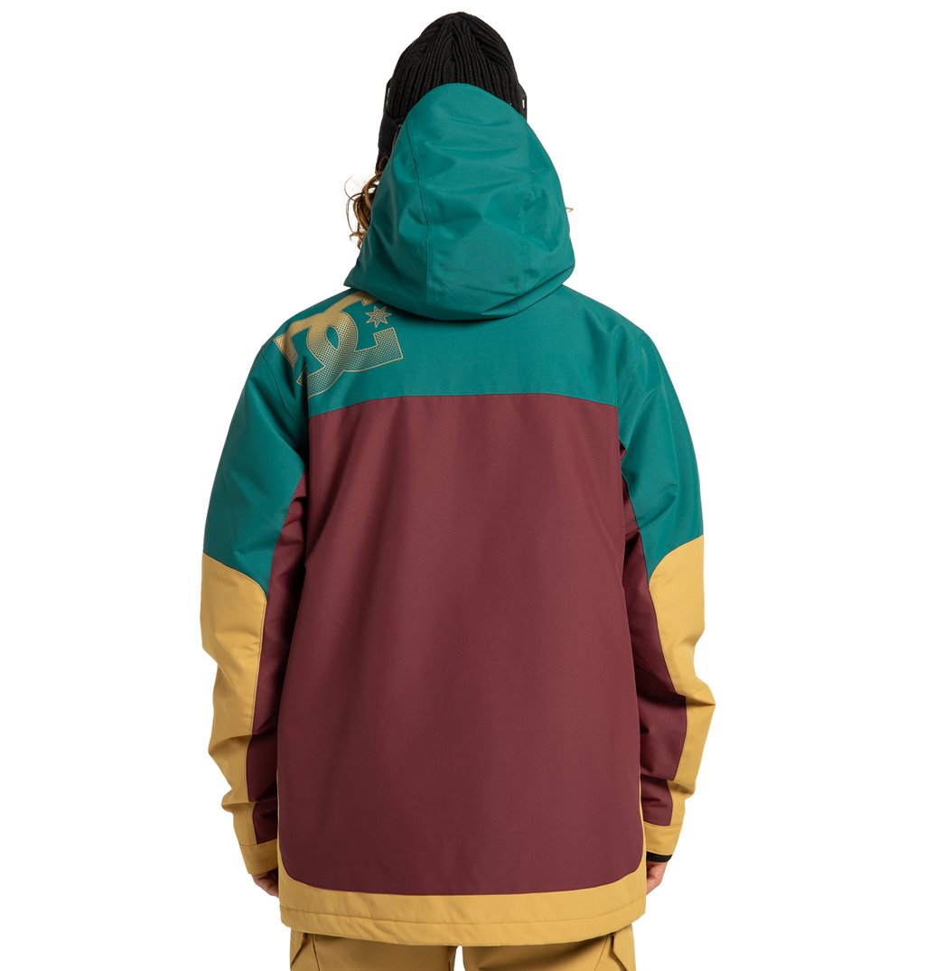 A person shown from the back is wearing the DC Shoes Men’s Defiant Insulated Jacket, a color-blocked winter adventure style in teal, burgundy, and yellow with a hood and a beanie. A large DC Shoes logo appears on the upper left shoulder of the jacket.