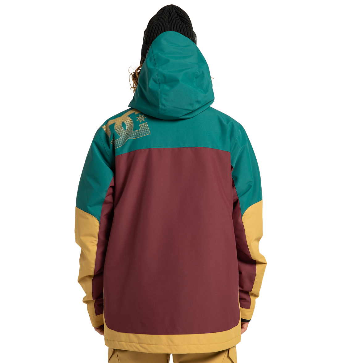 A person shown from the back is wearing the DC Shoes Men’s Defiant Insulated Jacket, a color-blocked winter adventure style in teal, burgundy, and yellow with a hood and a beanie. A large DC Shoes logo appears on the upper left shoulder of the jacket.