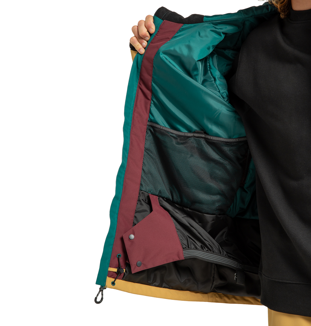 A person holds open the DC Shoes Men’s Defiant Insulated Jacket, revealing its teal, burgundy, and yellow interior with a mesh pocket and snap buttons. The person is wearing a black sweatshirt underneath.