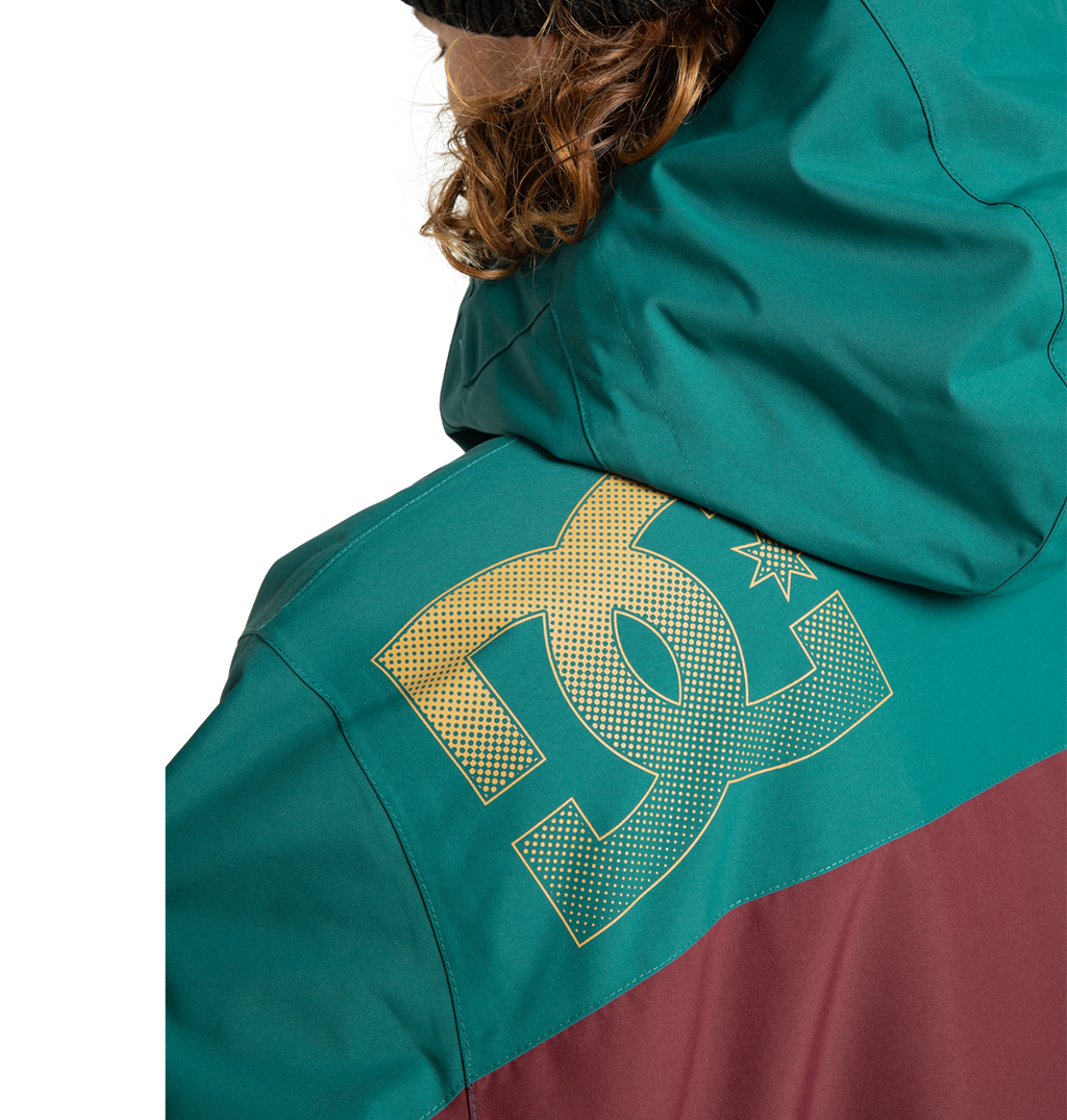 A person with brown curly hair wears the DC Shoes Men’s Defiant Insulated Jacket in teal and maroon, featuring a large yellow DC logo on the back shoulder. The hood is up, partially covering their head.