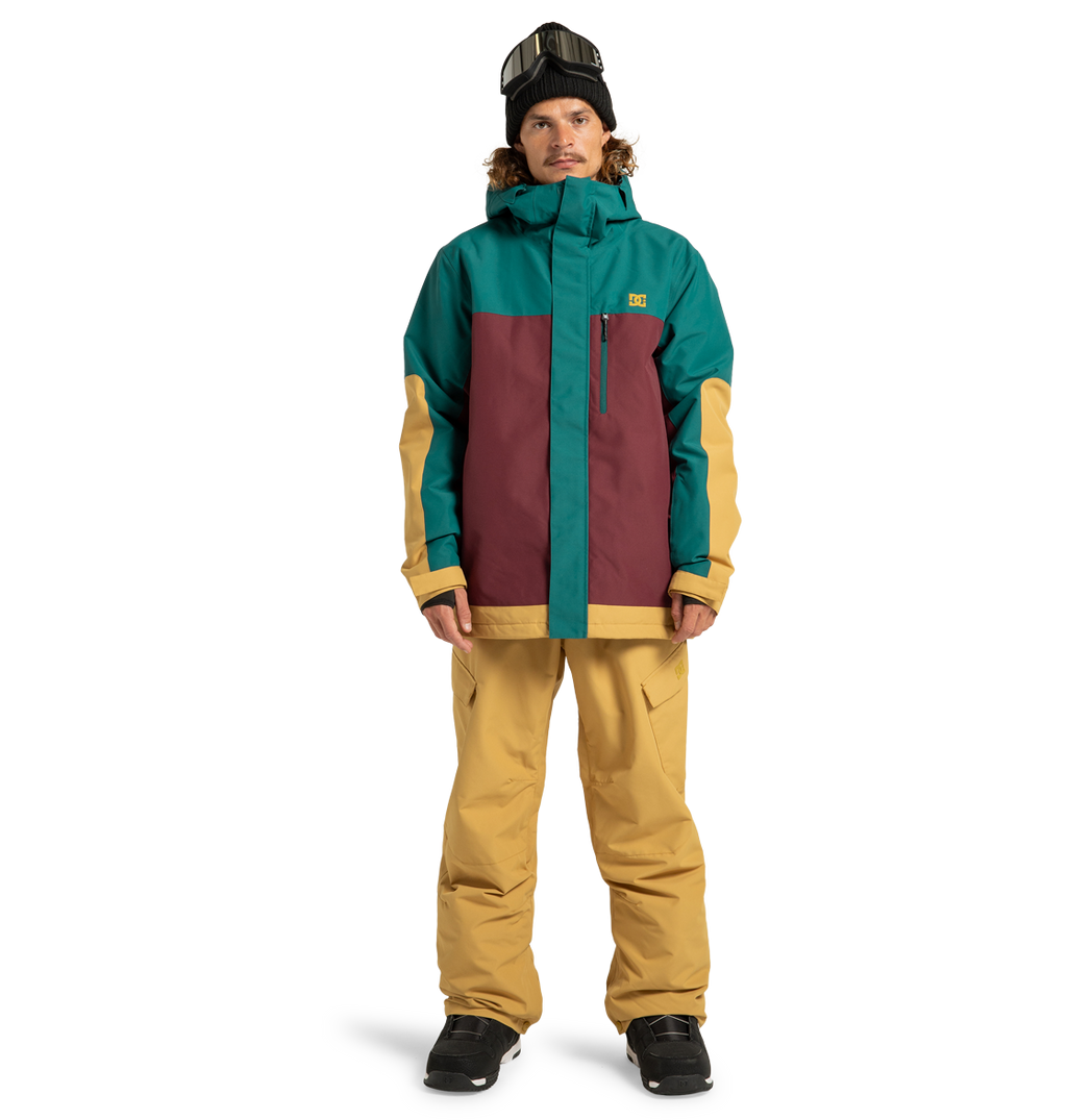A person wearing the DC Shoes Men’s Defiant Insulated Jacket in teal, burgundy, and tan, styled with tan snow pants, black gloves, black boots, and ski goggles on their head stands against a plain white background.