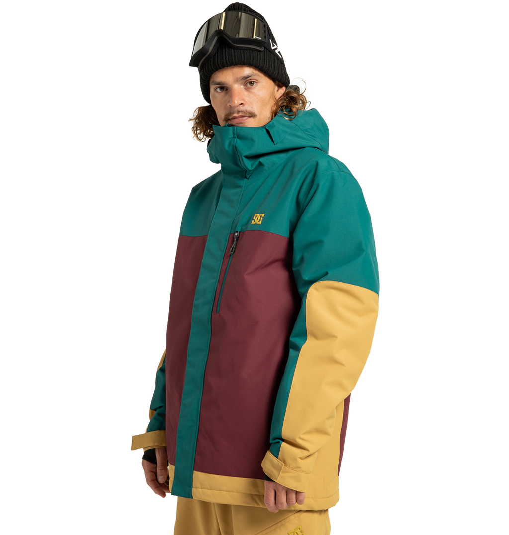A man wearing the DC Shoes Men’s Defiant Insulated Jacket in teal, maroon, and yellow—crafted from recycled polyester—paired with matching yellow pants and a black beanie topped with ski goggles, stands against a plain white background, ready for any winter adventure.
