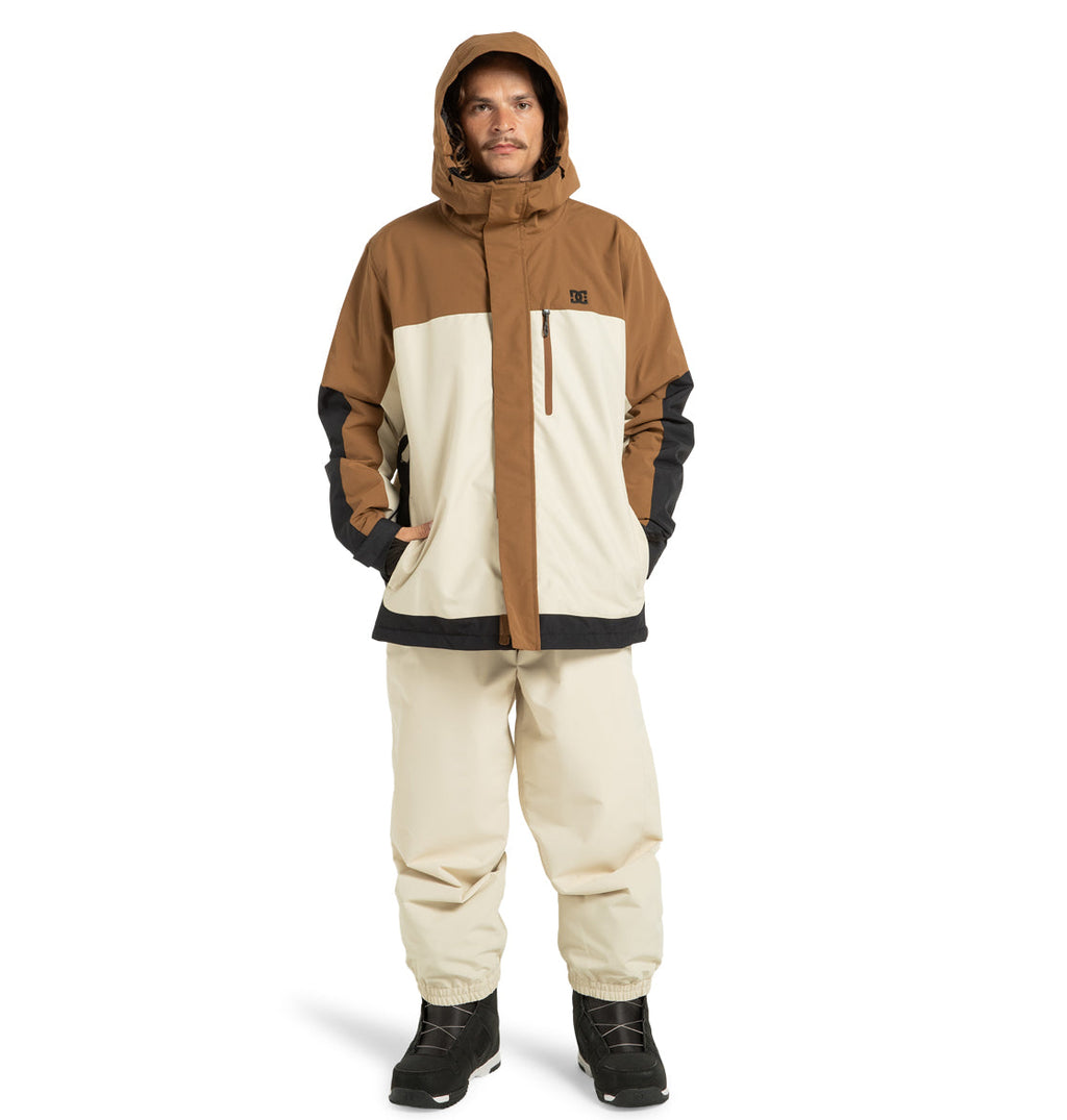 A person stands facing forward against a plain white background with hands in the pockets of a brown and cream DC Shoes Men’s Defiant Insulated Snow Jacket, paired with matching cream pants and black boots.