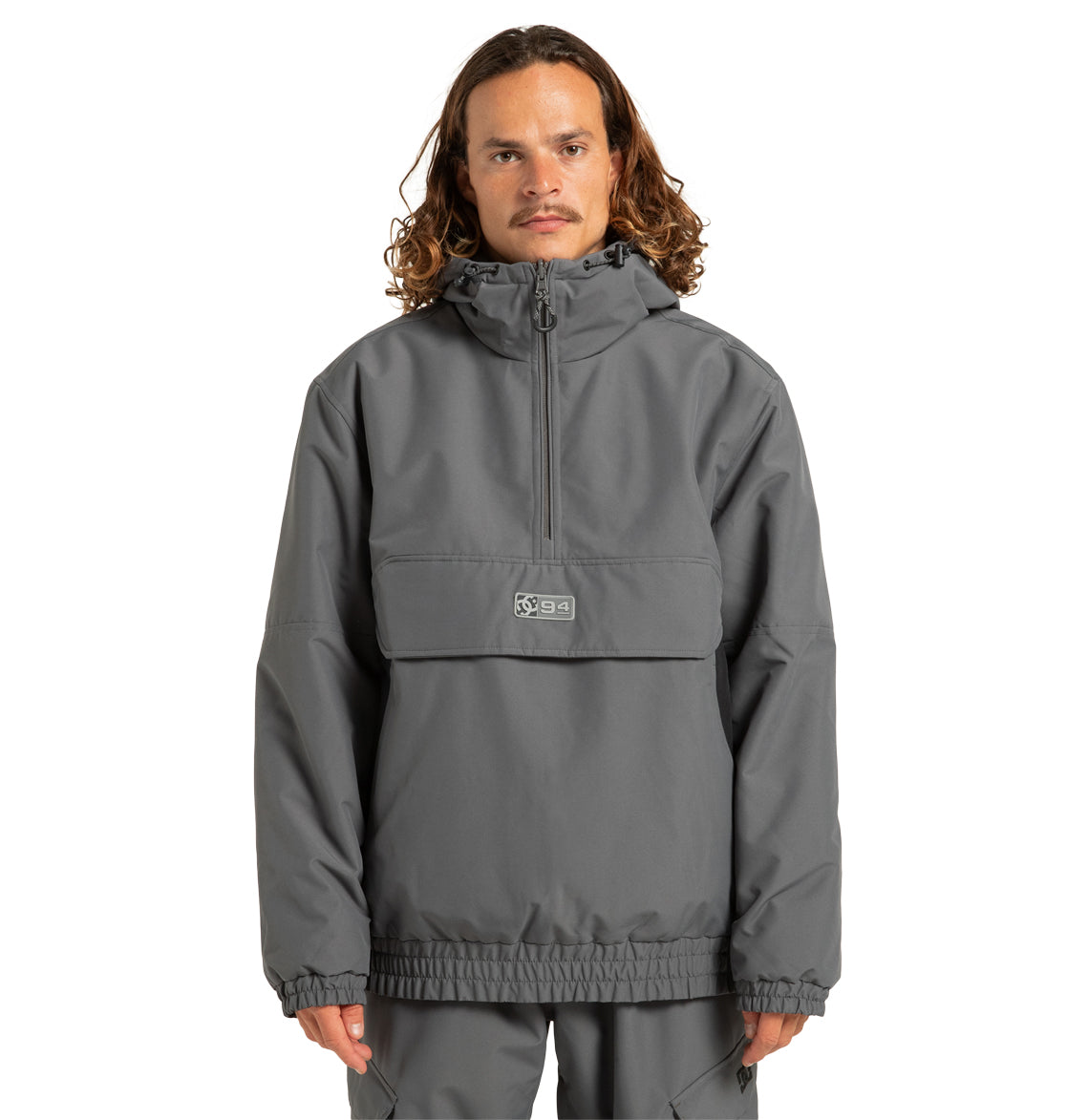 A person with wavy, shoulder-length hair wears the DC Shoes Unisex Nexus Reversible Insulated Anorak Jacket in gray—featuring a weather-resistant shell, half-zip design, front flap pocket, and elastic cuffs—standing against a plain white background.