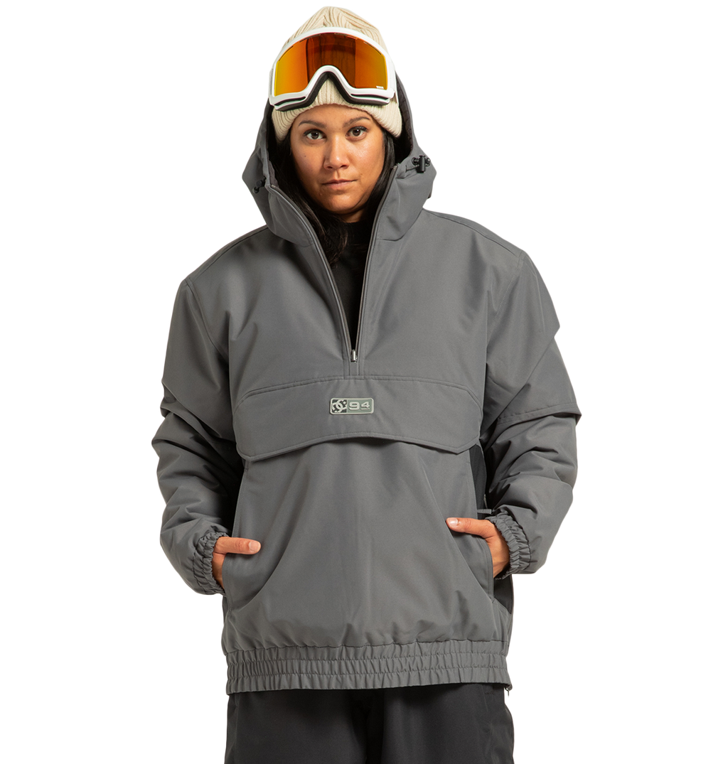 A person wearing the DC Shoes Unisex Nexus Reversible Insulated Anorak Jacket in gray, paired with a tan beanie and ski goggles, stands with hands in pockets against a plain white background.