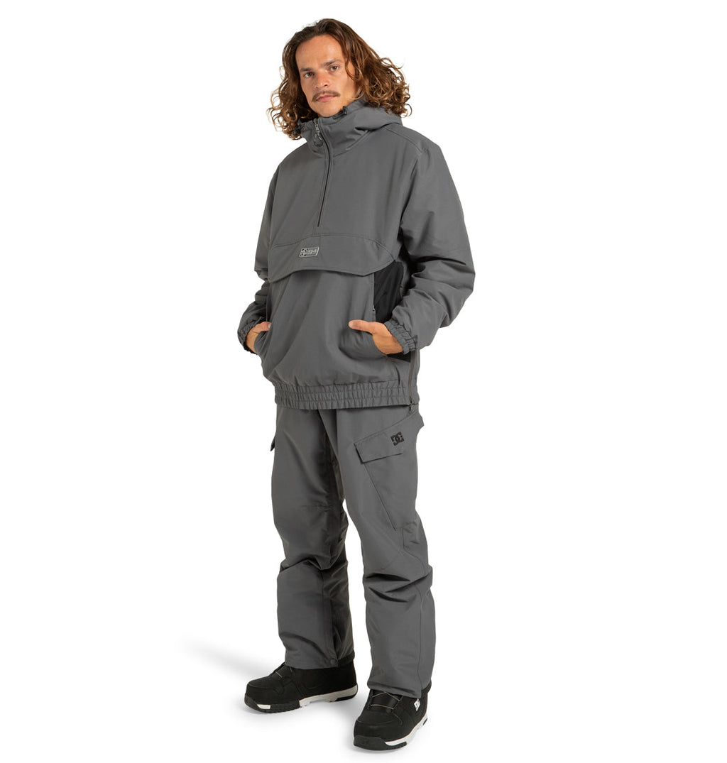 A man with long hair, hands in his pockets, stands facing slightly left wearing the DC Shoes Unisex Nexus Reversible Insulated Anorak Jacket with matching pants. He sports black sneakers and maintains a neutral expression.