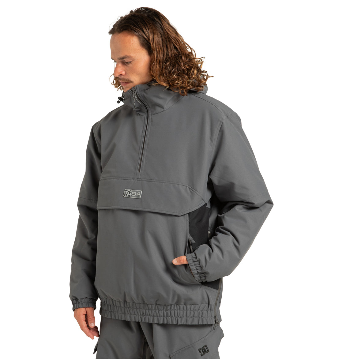 A person with long curly hair wears the loose-fitting, gray Unisex Nexus Reversible Insulated Anorak Jacket by DC Shoes, featuring a front zipper and hand pocket. The individual is looking down while standing against a plain white background.