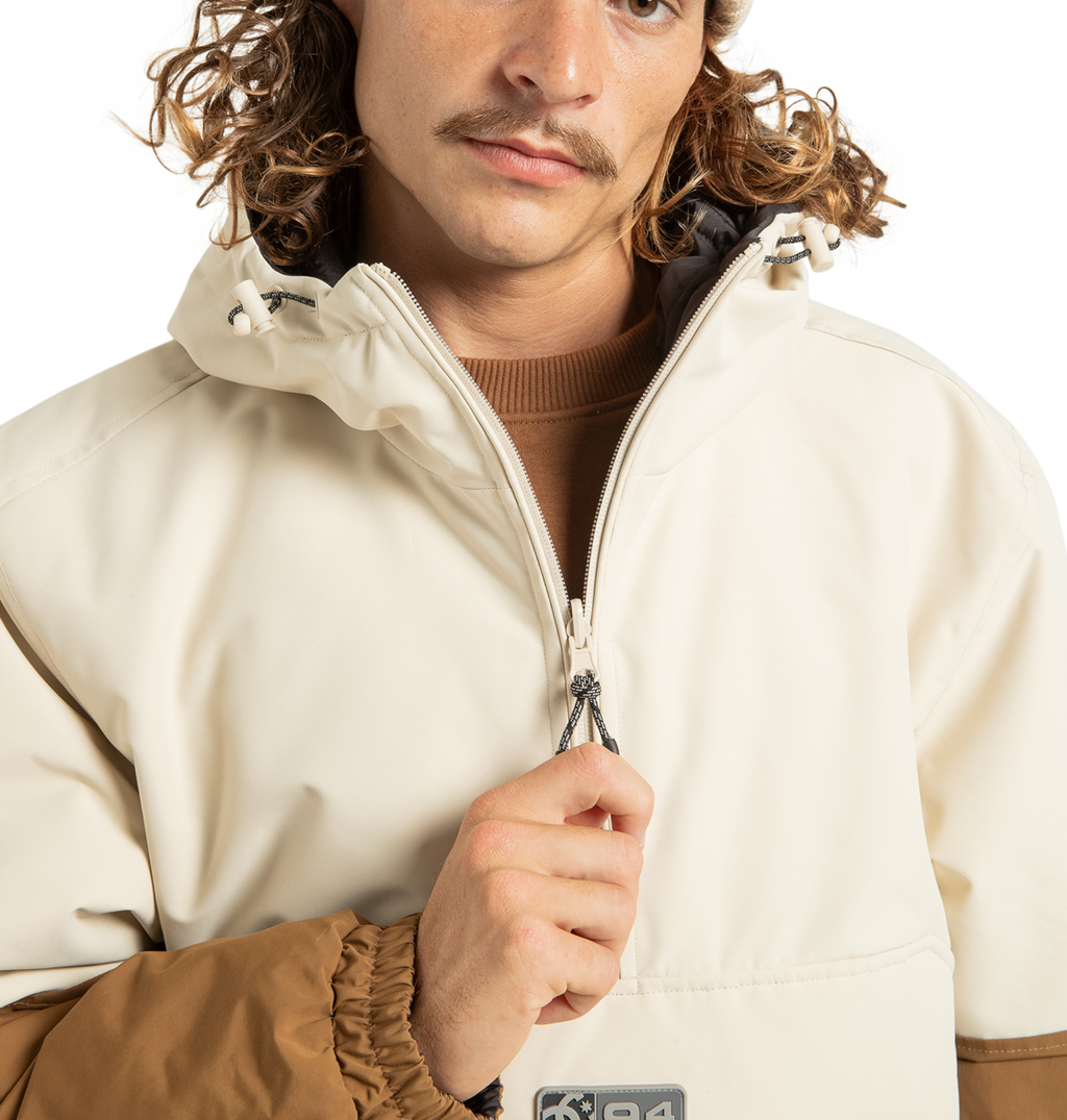 A person with wavy hair and a mustache wears the light-toned DC Shoes Unisex Nexus Reversible Insulated Anorak Jacket, featuring contrasting brown sleeves and crafted from recycled polyester. The zipper is partially up as the jacket stands out against a plain white background.