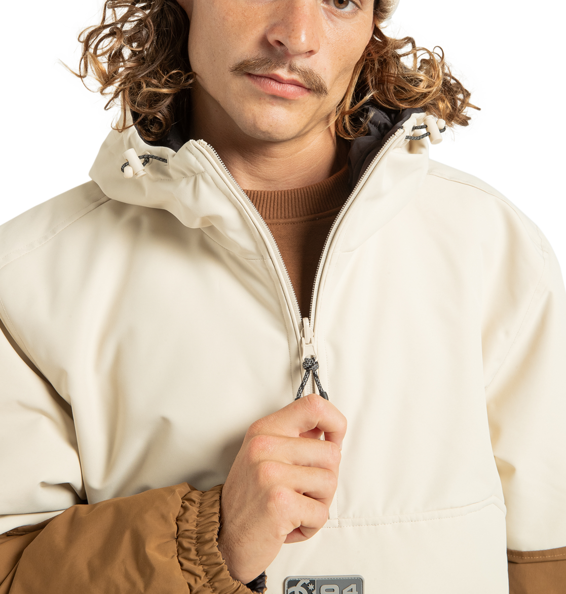 A person with wavy hair and a mustache wears the light-toned DC Shoes Unisex Nexus Reversible Insulated Anorak Jacket, featuring contrasting brown sleeves and crafted from recycled polyester. The zipper is partially up as the jacket stands out against a plain white background.
