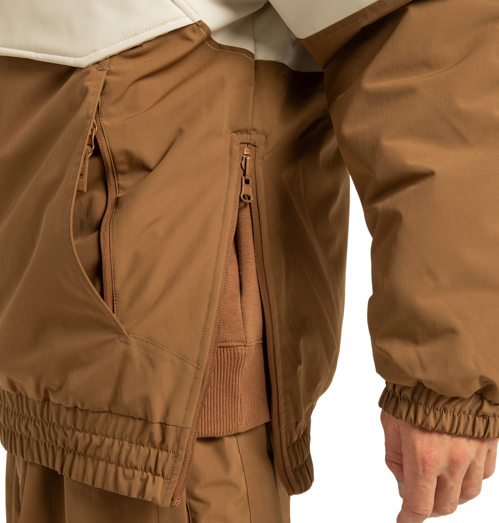 A close-up of a person wearing a brown DC Shoes Unisex Nexus Reversible Insulated Anorak Jacket with their hand resting at their side, showcasing the zippered pockets and elastic cuffs. This recycled polyester jacket is layered over other brown clothing, making it perfect for outdoor adventures.