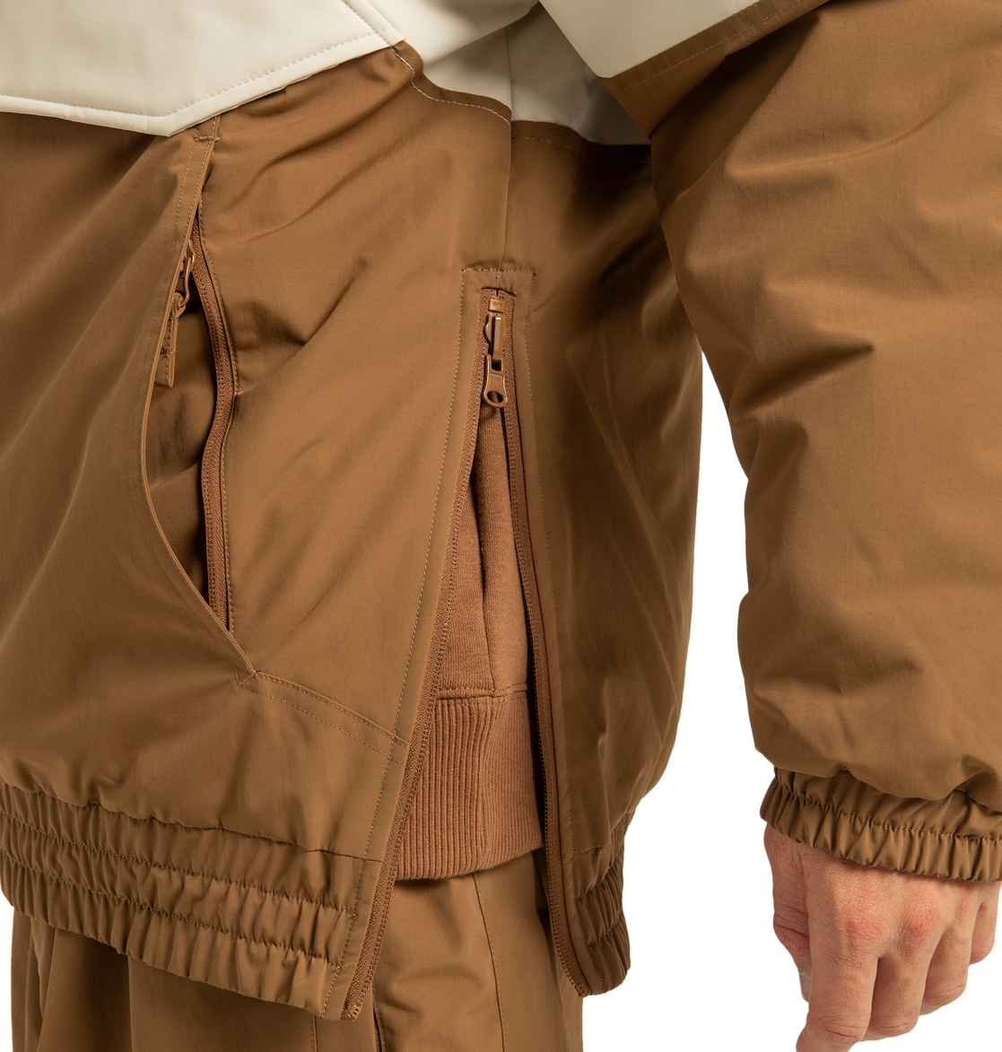 A close-up of a person wearing a brown DC Shoes Unisex Nexus Reversible Insulated Anorak Jacket with their hand resting at their side, showcasing the zippered pockets and elastic cuffs. This recycled polyester jacket is layered over other brown clothing, making it perfect for outdoor adventures.