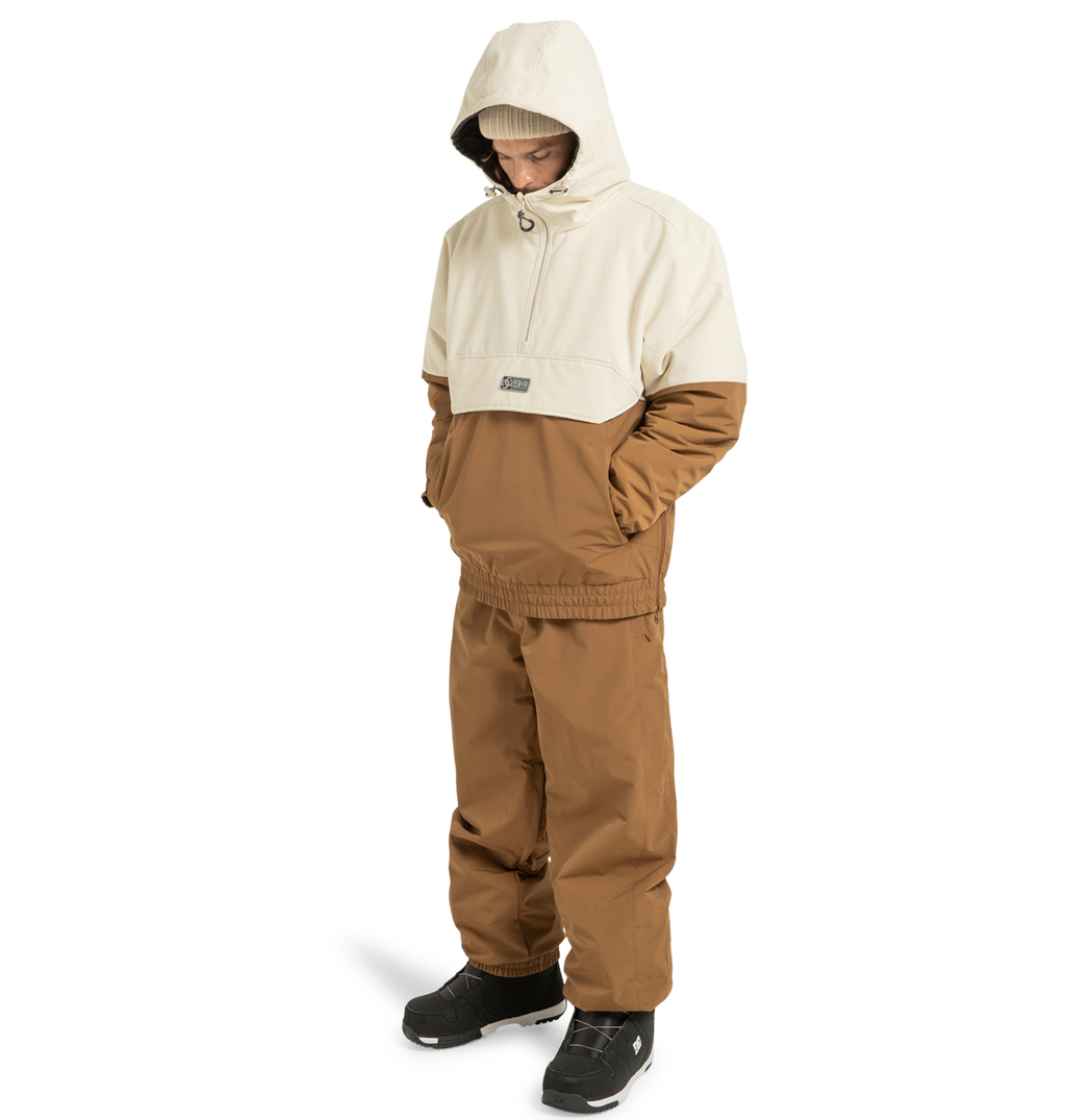 A person stands against a plain white background wearing the DC Shoes Unisex Nexus Reversible Insulated Anorak Jacket in two-tone brown and beige, crafted from recycled polyester. They pair it with matching brown pants, black boots, and a beige beanie, hands in pockets—perfectly outfitted for any outdoor adventure.
