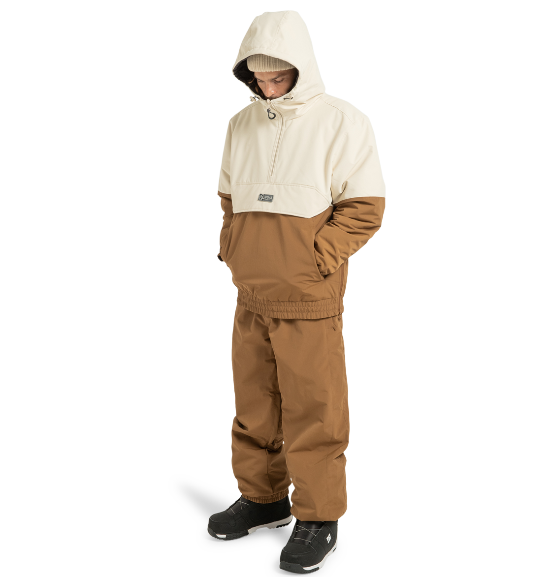 A person stands against a plain white background wearing the DC Shoes Unisex Nexus Reversible Insulated Anorak Jacket in two-tone brown and beige, crafted from recycled polyester. They pair it with matching brown pants, black boots, and a beige beanie, hands in pockets—perfectly outfitted for any outdoor adventure.
