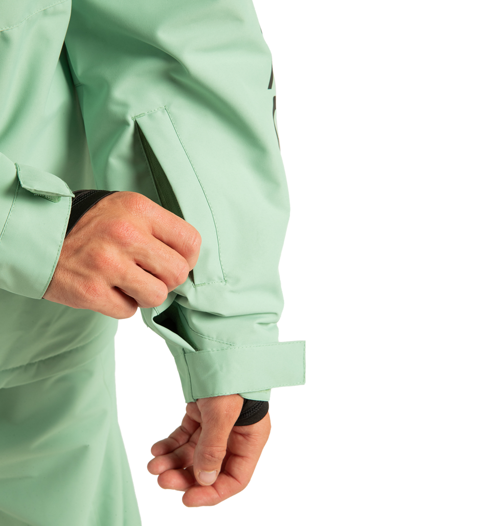 A person unzips a sleeve pocket on the DC Shoes Unisex Basis Insulated Jacket in light green, crafted from recycled polyester, with a Velcro strap secured at the wrist—ideal for winter adventures.
