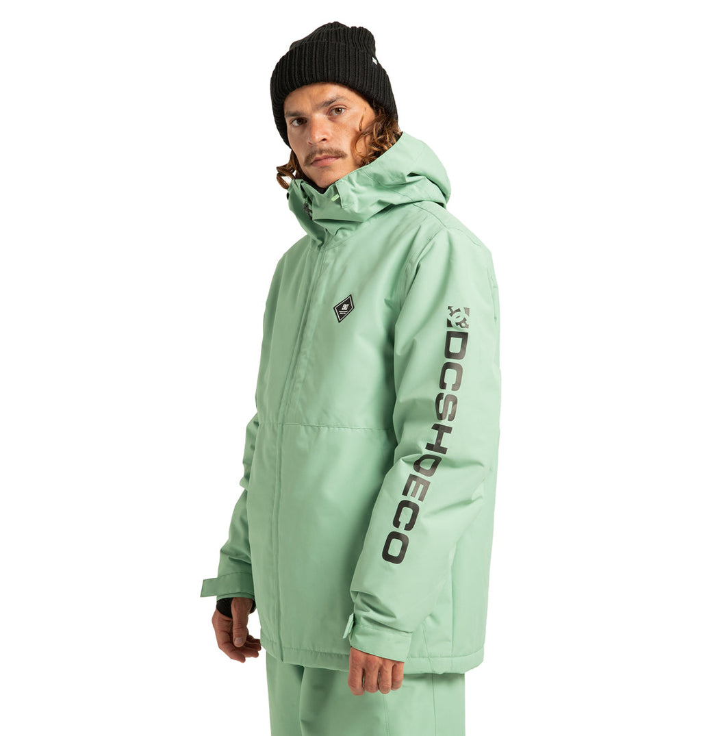 A person wearing the DC Shoes Unisex Basis Insulated Jacket in mint green, paired with matching pants and a black beanie, perfect for winter activities. Made from recycled polyester, the jacket features “DCSHOECO” written in black on the left sleeve. The person faces the camera with a neutral expression.
