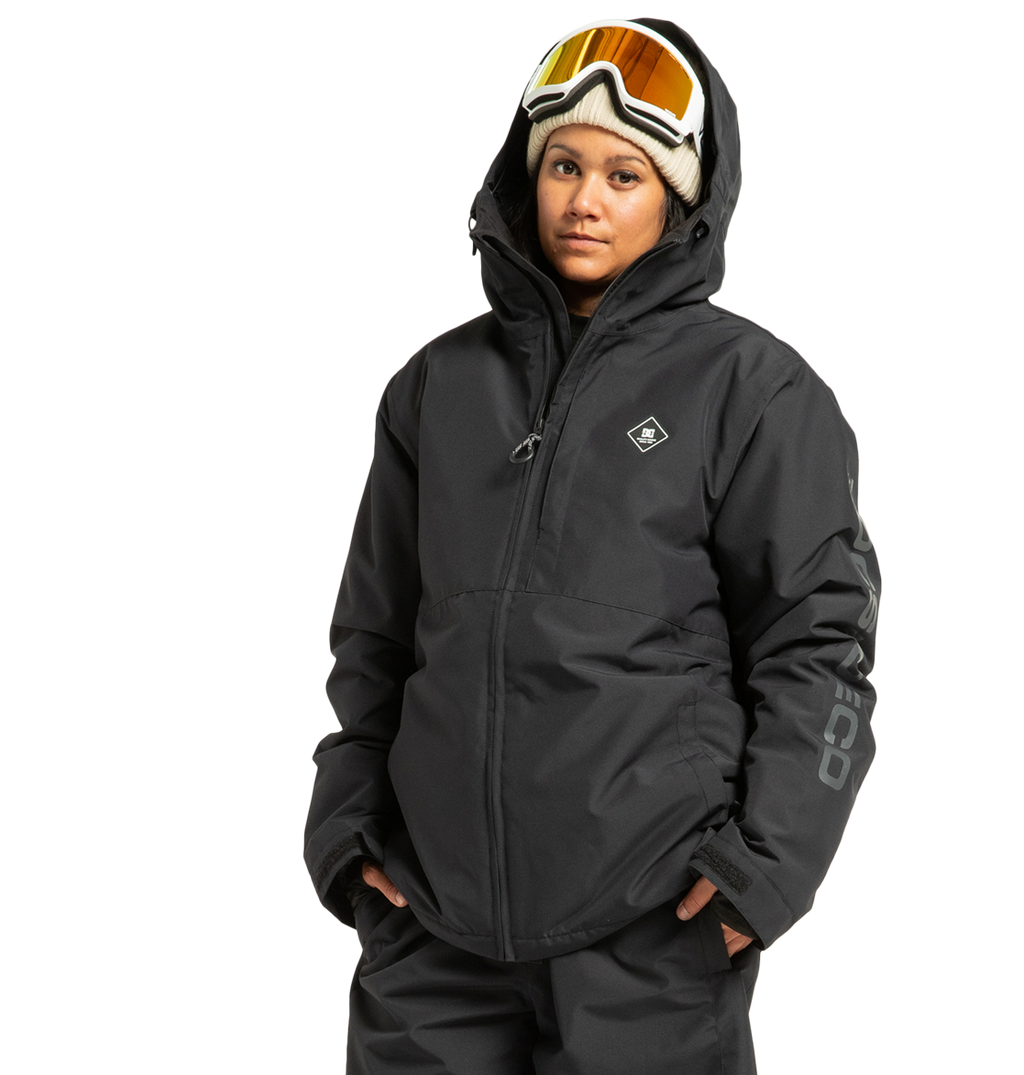 Wearing the DC Shoes Unisex Basis Insulated Jacket, along with black snow pants, ski goggles, and a beige beanie, a person stands with hands in pockets against a plain white background.