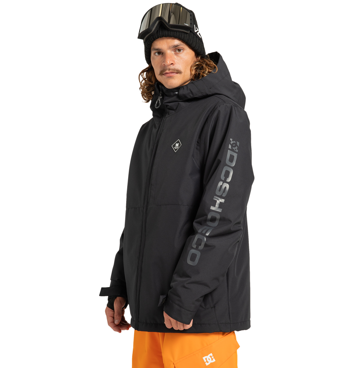 A person wearing the Unisex Basis Insulated Jacket by DC Shoes—a black, insulated outdoor jacket crafted from recycled polyester with “DCSHOECO” branding on the sleeve—paired with orange pants, ski goggles on their head, and a black beanie, stands against a plain white background.