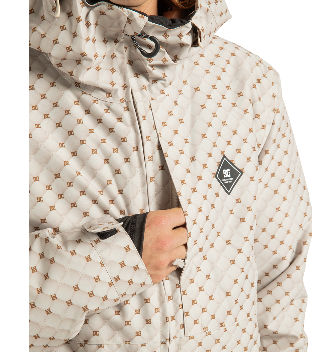 A person wearing the DC Shoes Unisex Basis Print Insulated Jacket, a light-colored patterned outerwear piece with a diamond-shaped logo on the chest, partially unzipped to reveal a black inner layer. Crafted from recycled polyester, this jacket features a high collar and adjustable cuffs.