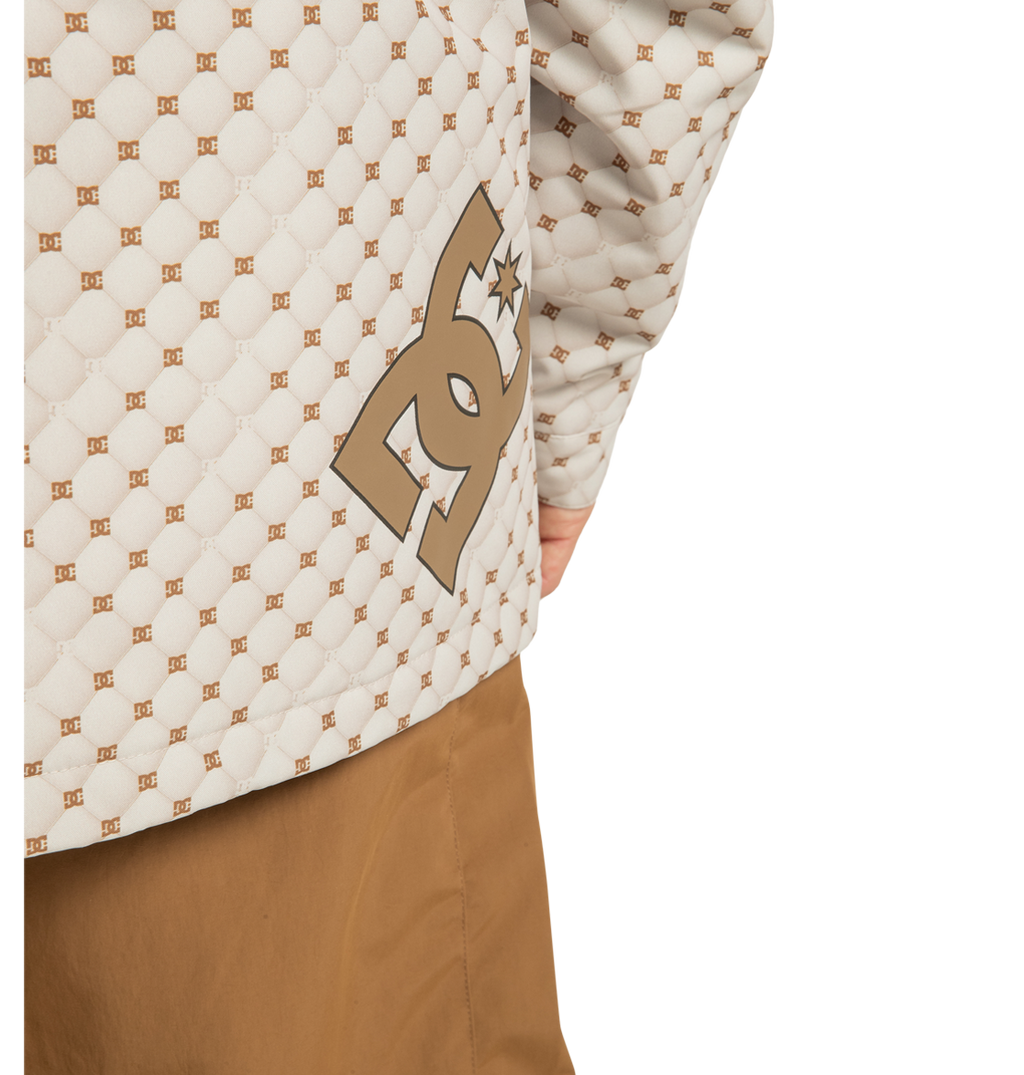 Close-up of a person wearing the Unisex Basis Print Insulated Jacket by DC Shoes, featuring a beige quilted design with a brown repeating logo pattern and prominent DC branding, crafted from recycled polyester, paired with brown pants. Only part of the torso and right arm are visible.