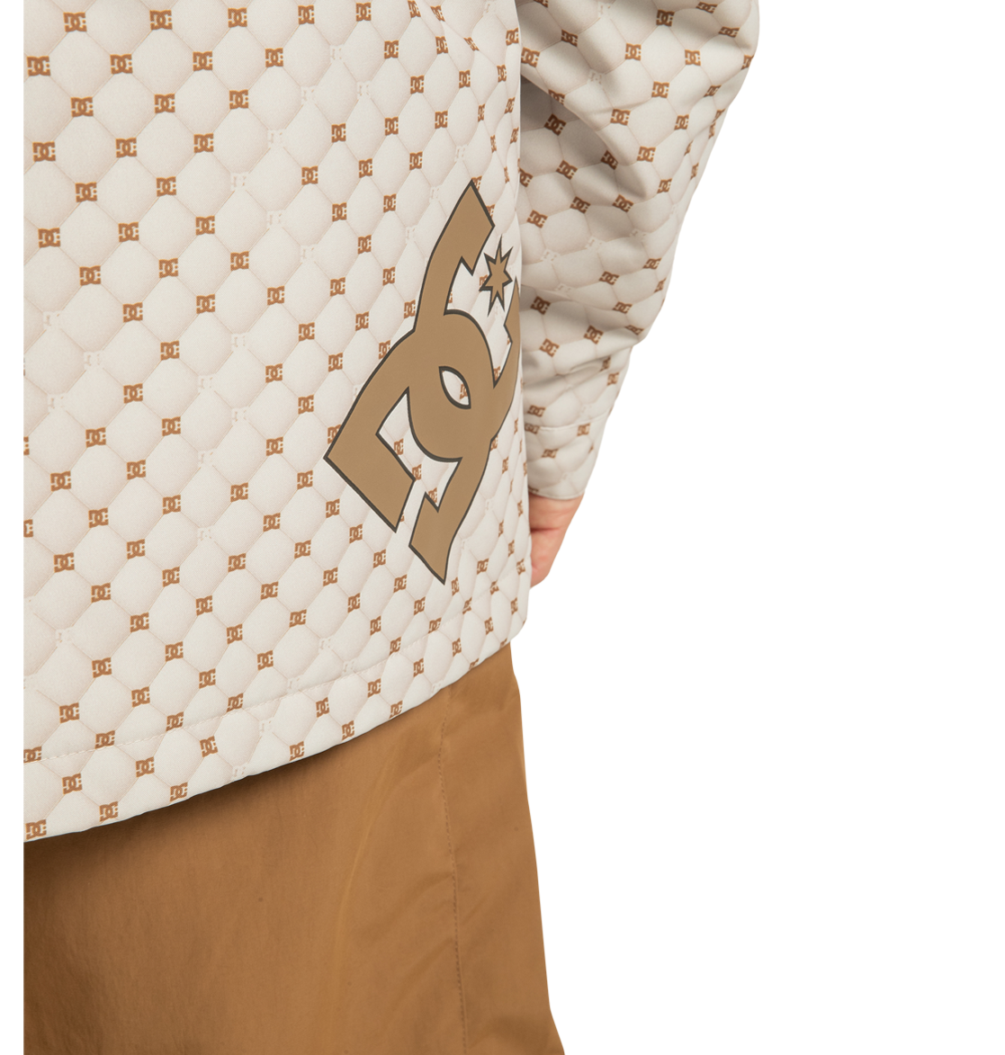 Close-up of a person wearing the Unisex Basis Print Insulated Jacket by DC Shoes, featuring a beige quilted design with a brown repeating logo pattern and prominent DC branding, crafted from recycled polyester, paired with brown pants. Only part of the torso and right arm are visible.
