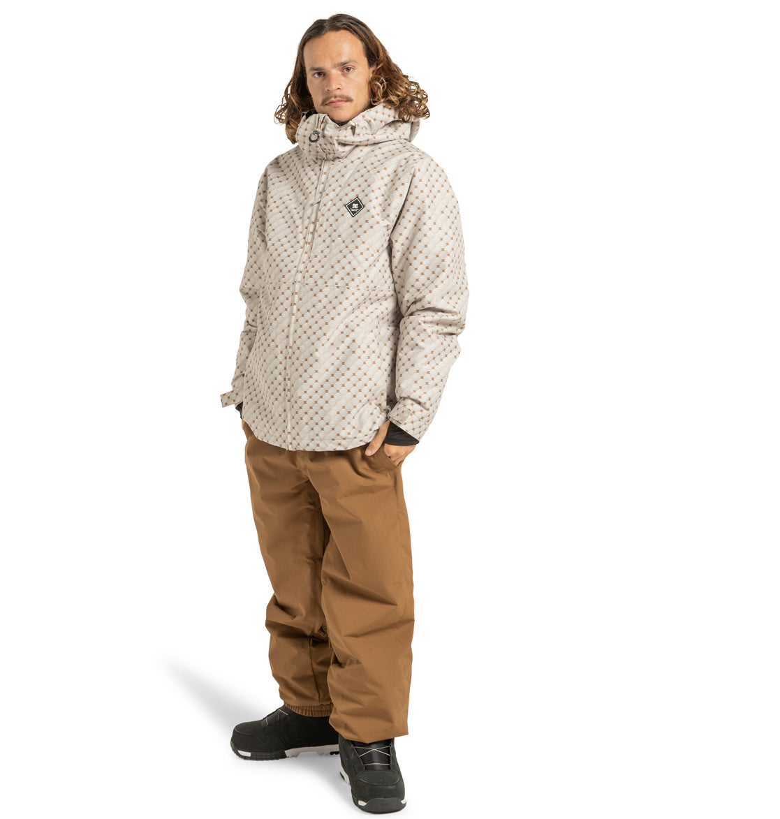A person with long hair stands against a white background wearing the DC Shoes Unisex Basis Print Insulated Jacket—featuring a light-colored pattern—paired with brown pants and black shoes.