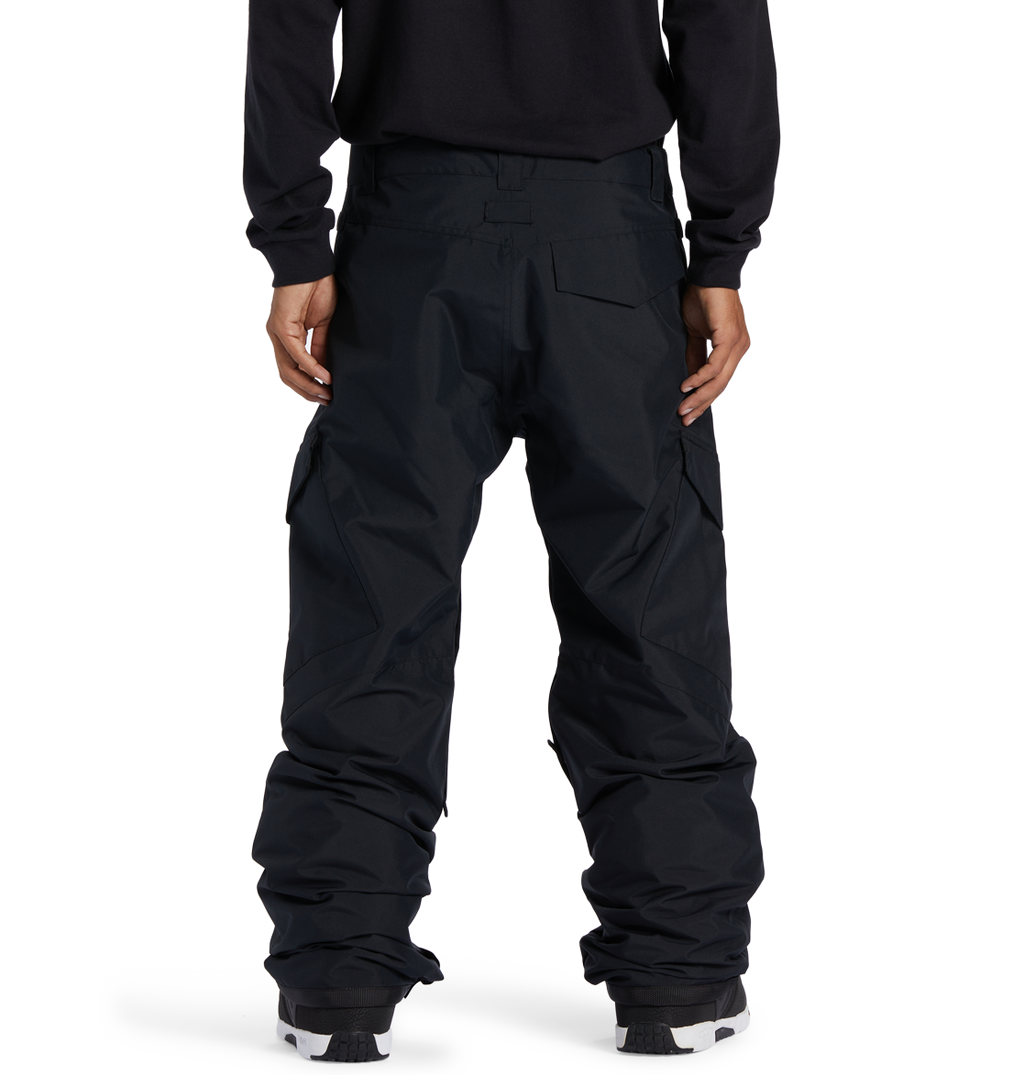 A man stands facing away, wearing DC Shoes Men’s Banshee Insulated Pants—loose-fitting black cargo pants designed for winter, featuring large pockets and covering the tops of his shoes—and a black long-sleeve shirt, with his hands relaxed at his sides.